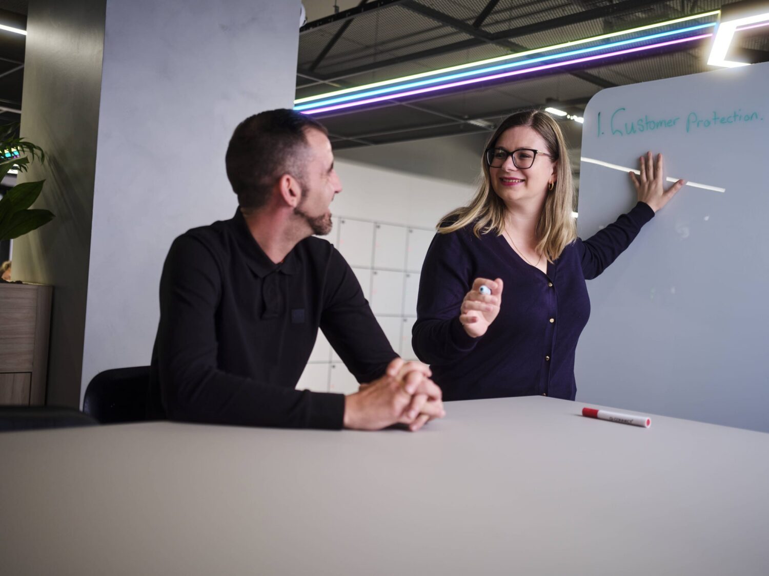 A man sits at a table while a woman points to a whiteboard during a discussion in a modern office.