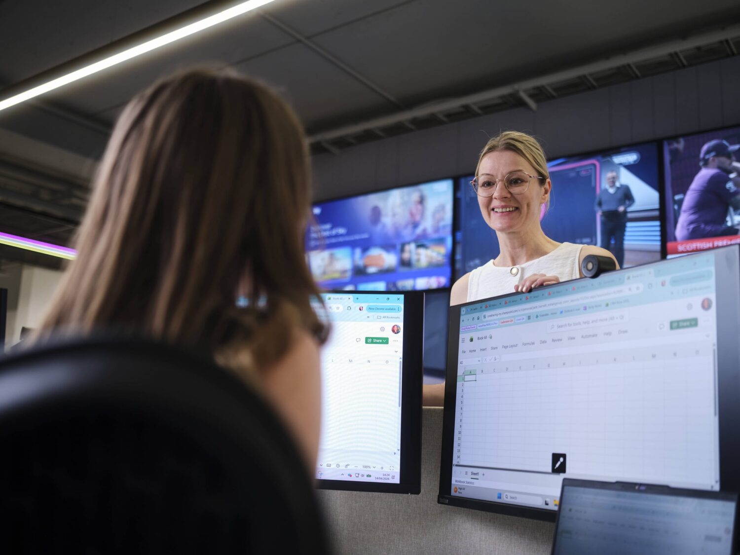 Smiling person stands beside a desk with several computer monitors in an office.