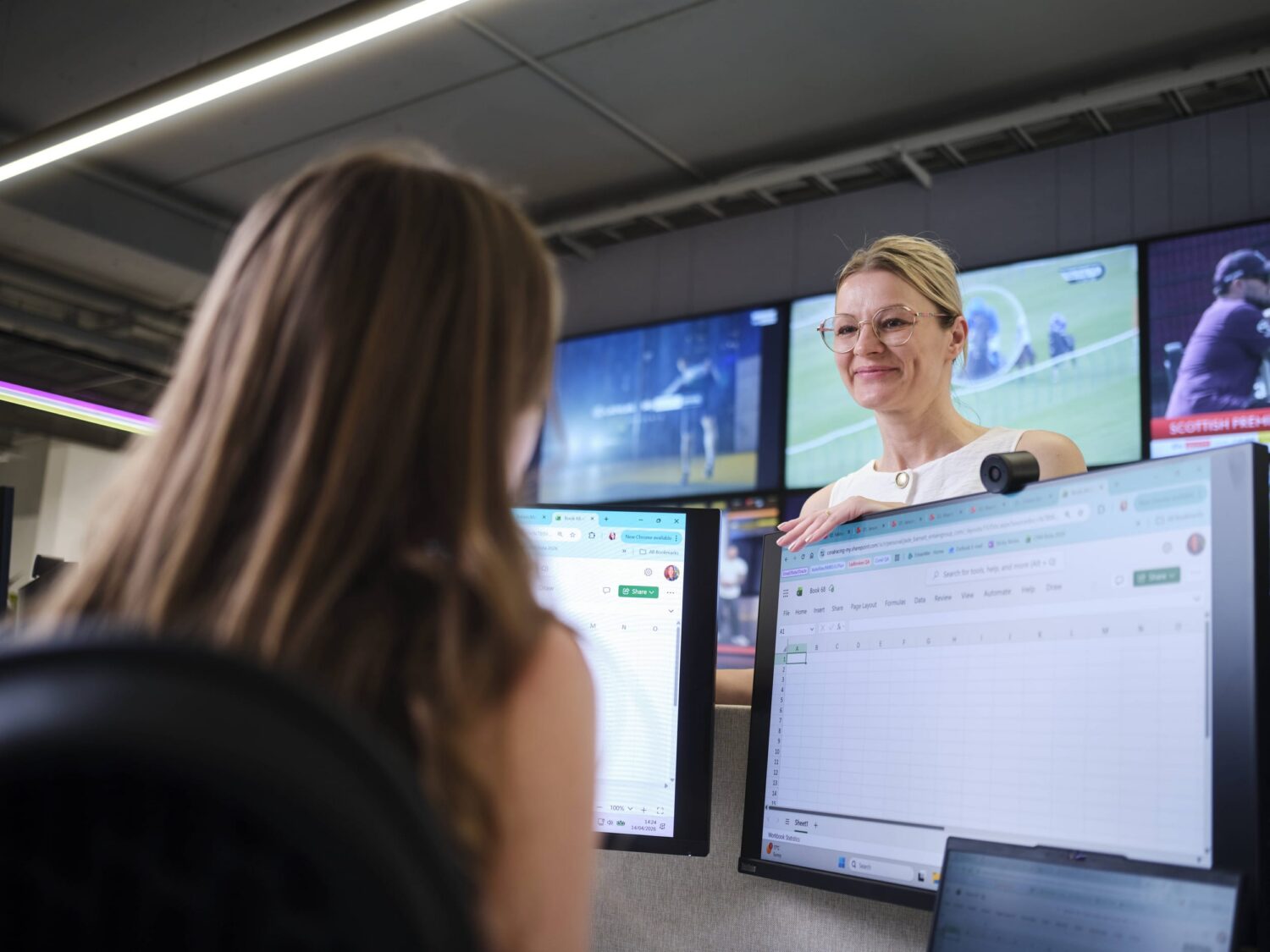 A blonde woman stands beside several computer monitors in an open office, smiling at a colleague seated with their back to the camera.