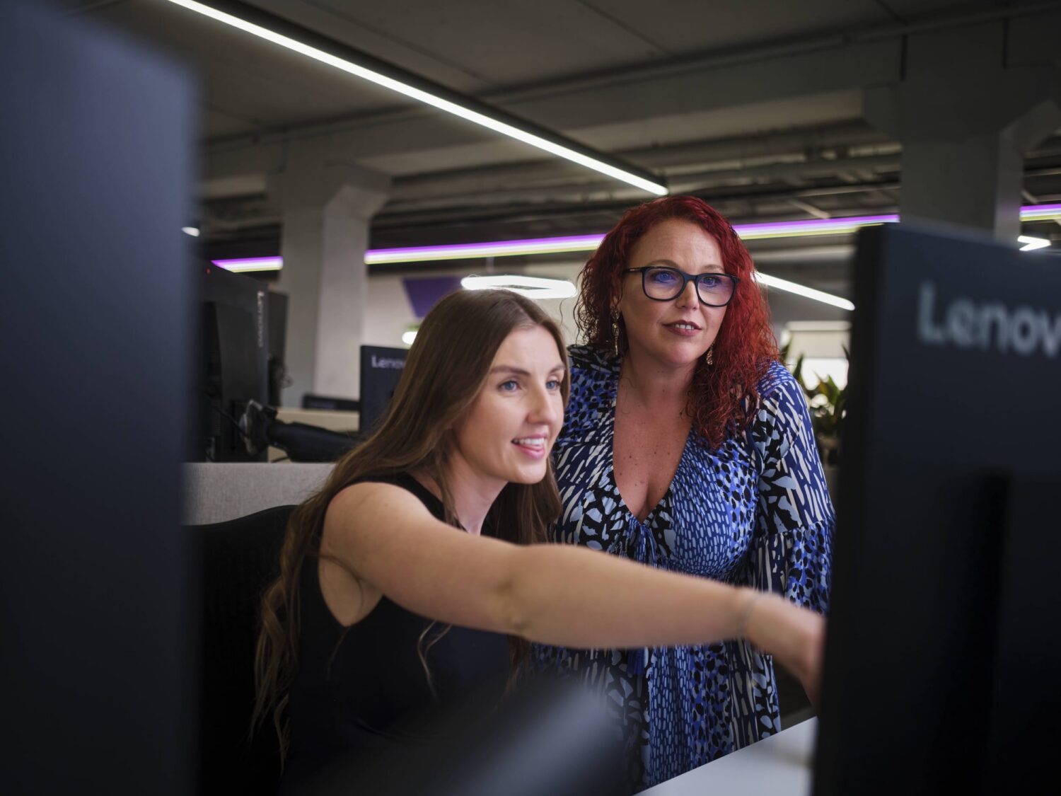 Two women collaborate at a computer in a Lenovo workstation area.