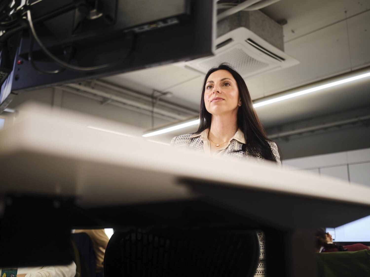 A woman seated at a desk in a modern office, looking upward toward a monitor mounted above.