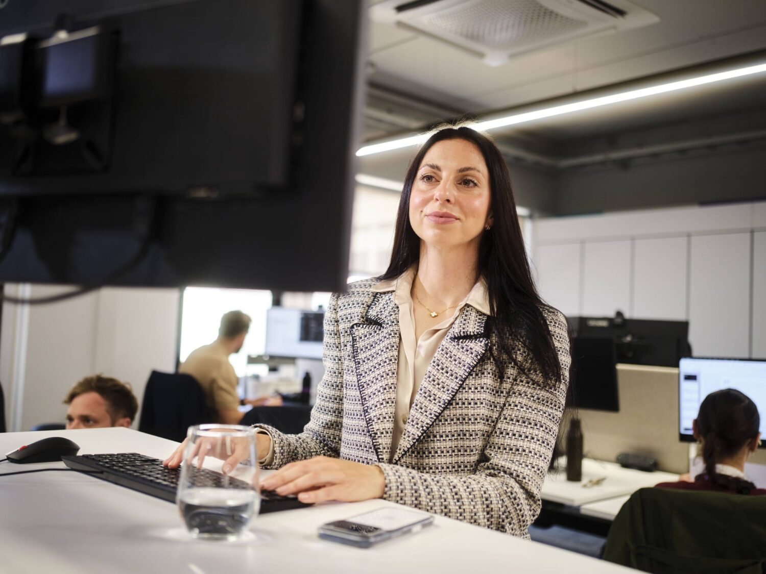 A businesswoman sits at her desk in an open-plan office, looking at her computer screen with a glass of water and papers on the desk.