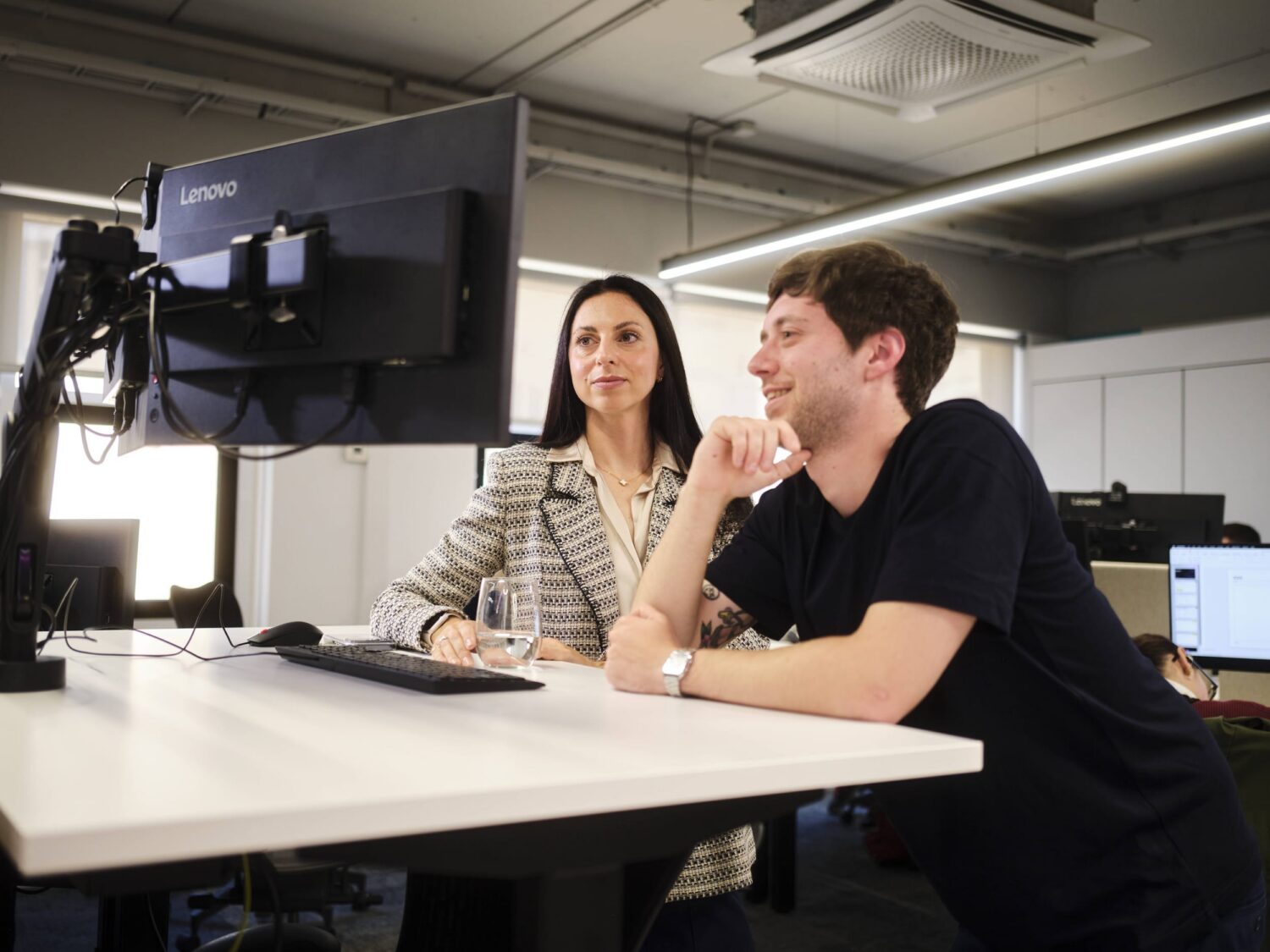 Two coworkers sit at a white desk in a modern studio, looking at a computer monitor beside camera equipment.