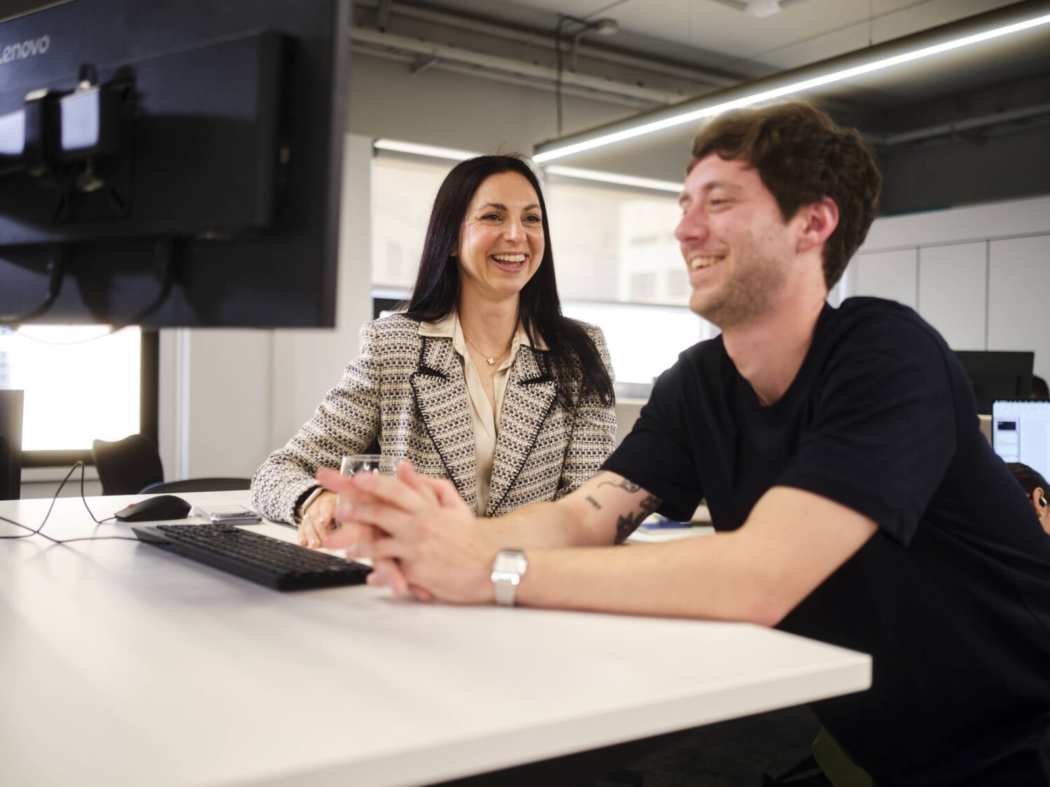 A woman and a man sit at a white desk in an office, smiling at each other.