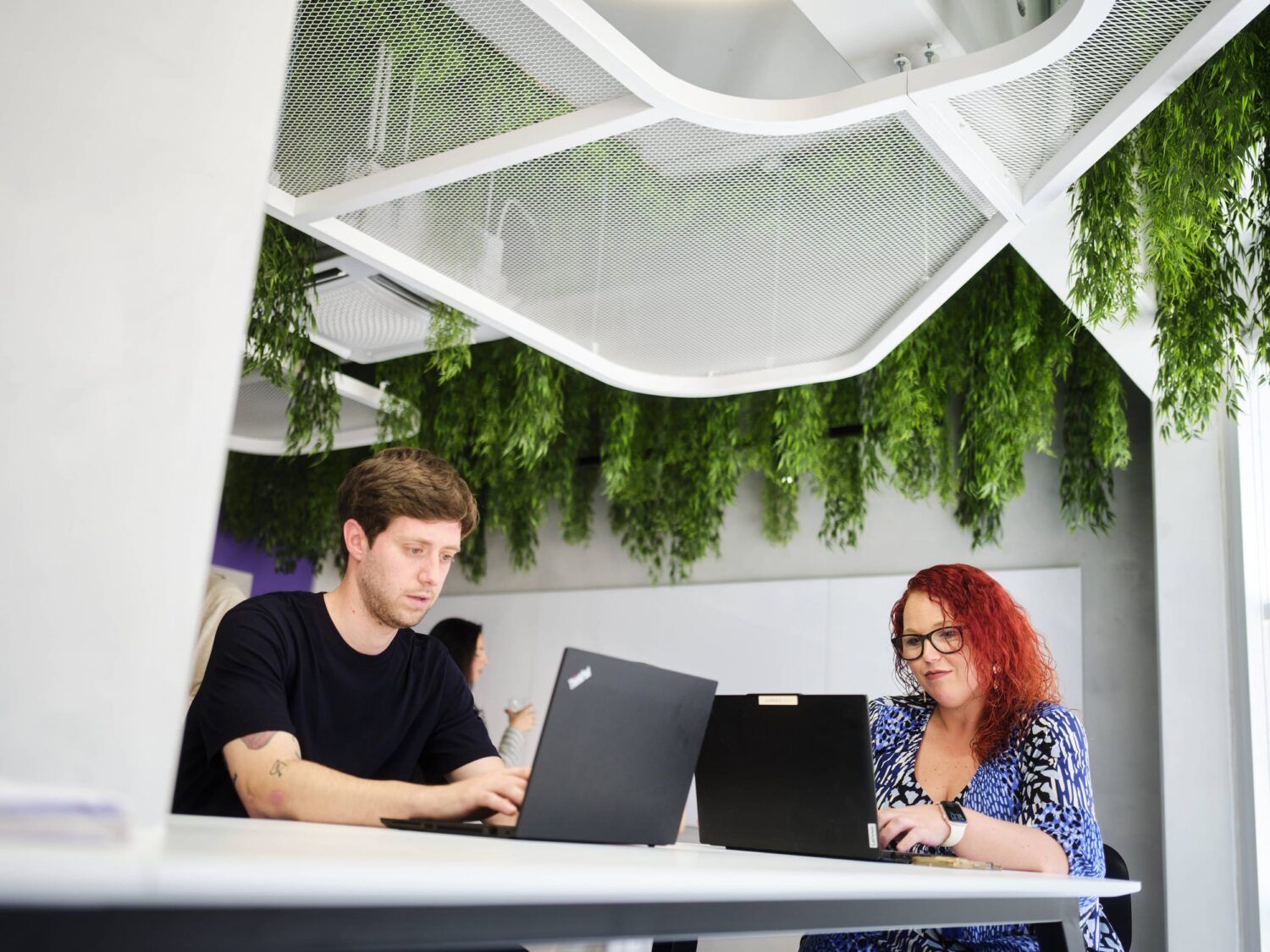 Two coworkers work on laptops at a white table in a bright office with hanging greenery.