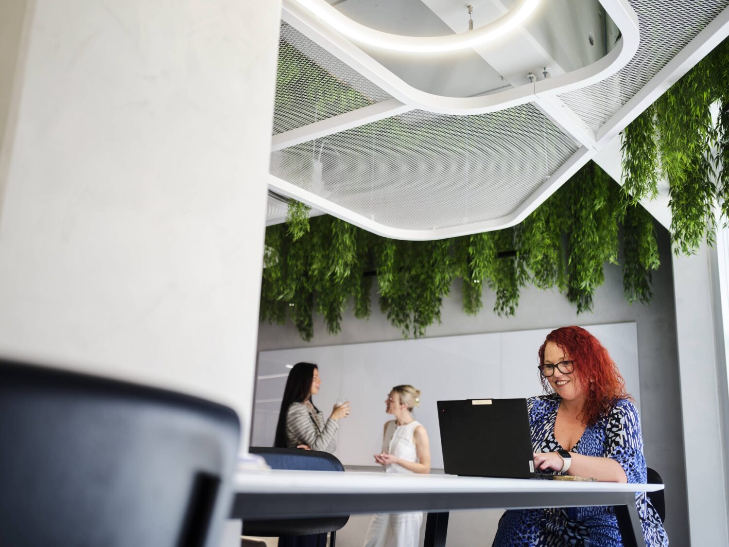 Three coworkers in a bright modern office with hanging greenery and a laptop on a table.
