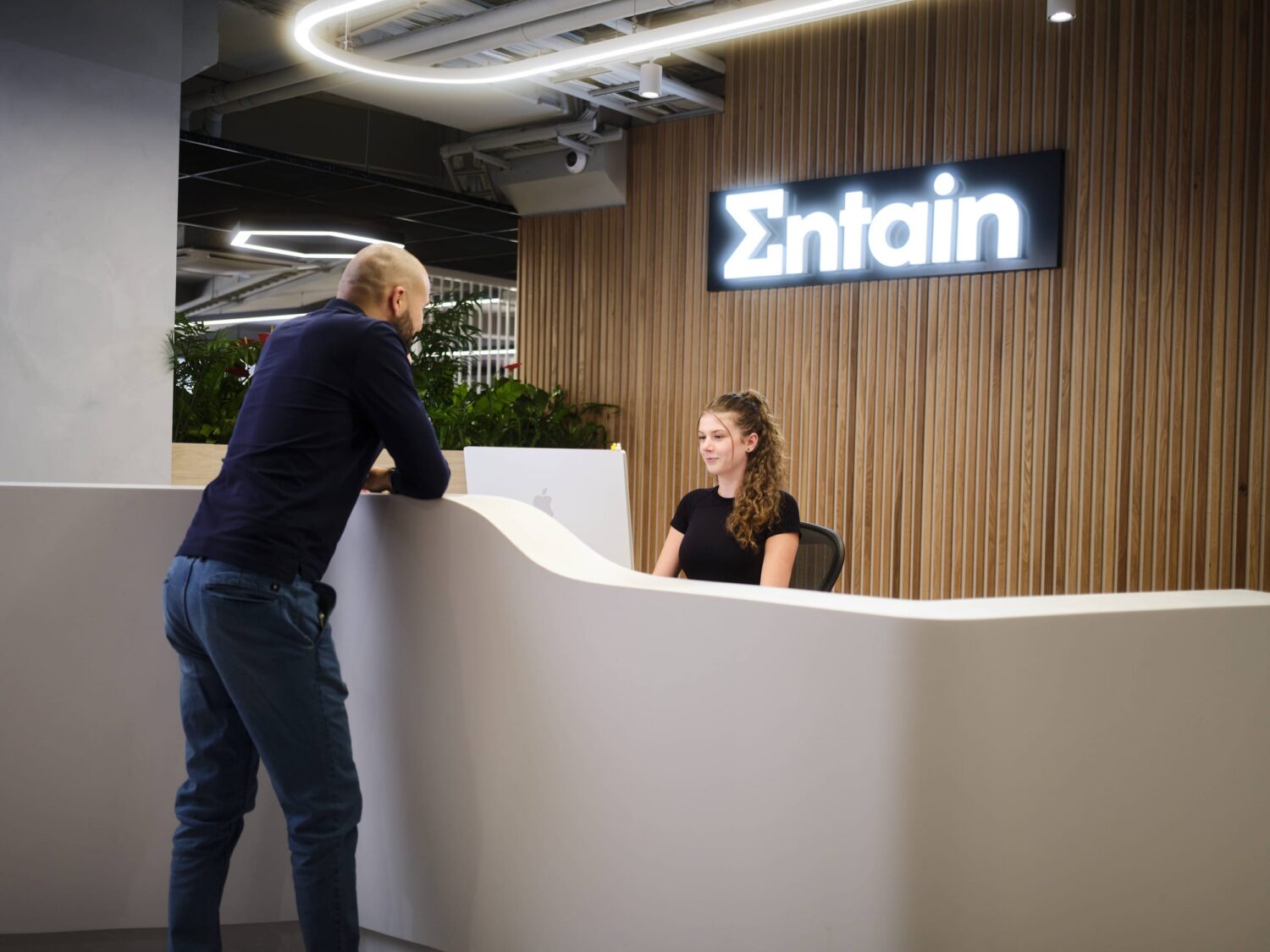 A man leans on a curved white reception desk while a woman sits behind it in a modern lobby with an Entain sign on a wood-paneled wall.