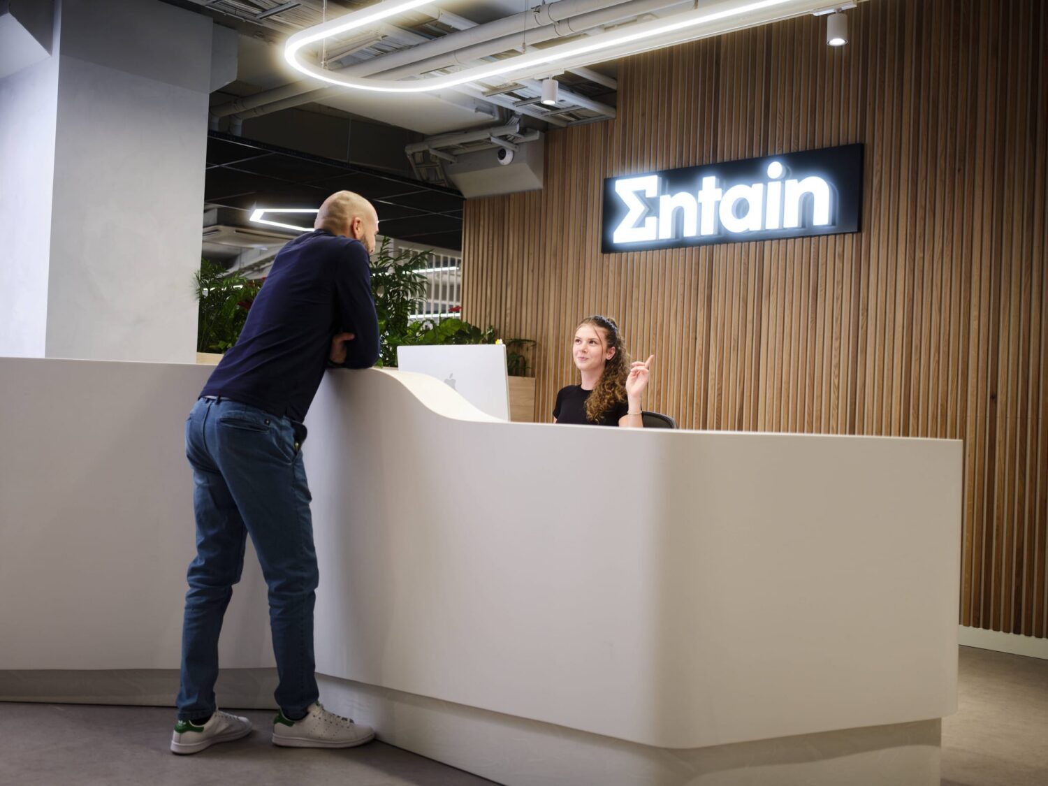 A man leans on a white reception desk while speaking to a receptionist in a modern office lobby with an illuminated Entain sign on a wood-paneled wall.
