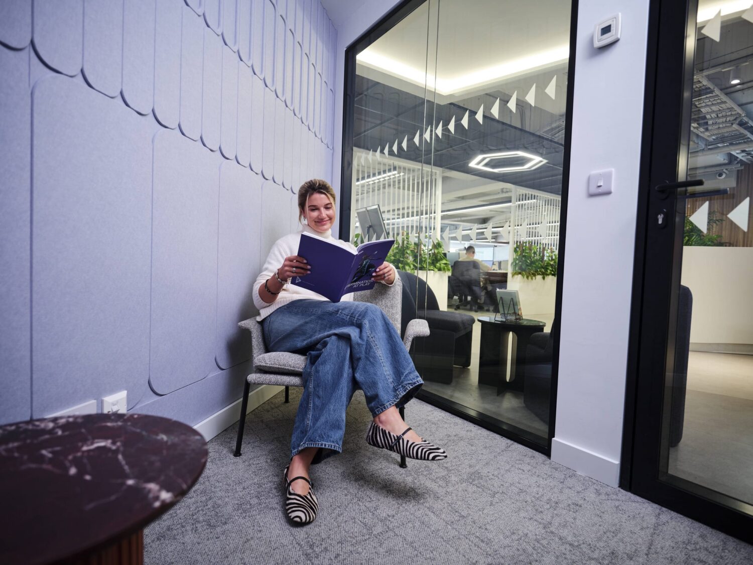 A person sits in a modern office lounge, reading a purple binder beside a glass door.