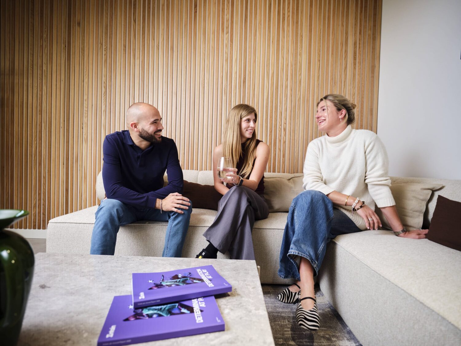 Three people sit on a light-gray sofa in a modern living room, chatting, with a purple magazine on the coffee table.