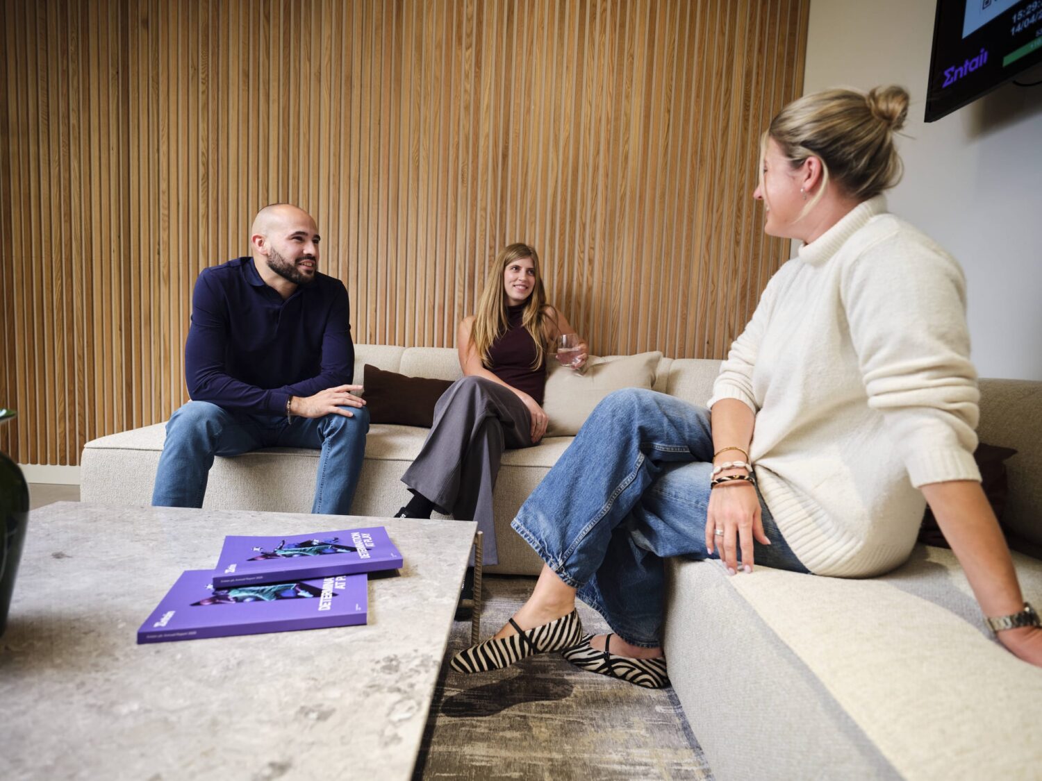 Three people sit on a beige sectional sofa in a living room, chatting, with a purple tray on the coffee table in front.