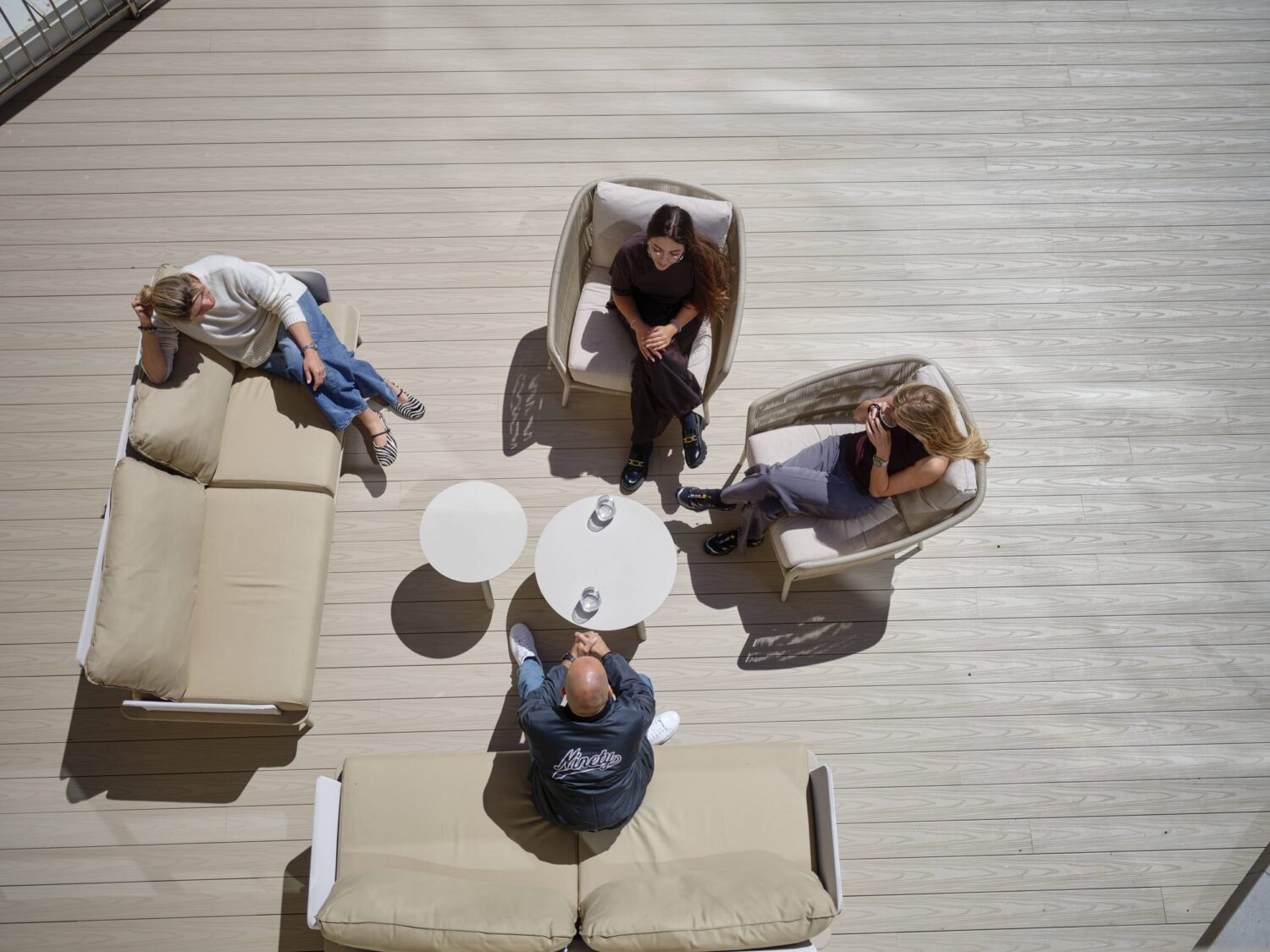 Overhead view of several people relaxing on beige outdoor couches arranged around a small white round table on a light wooden deck.