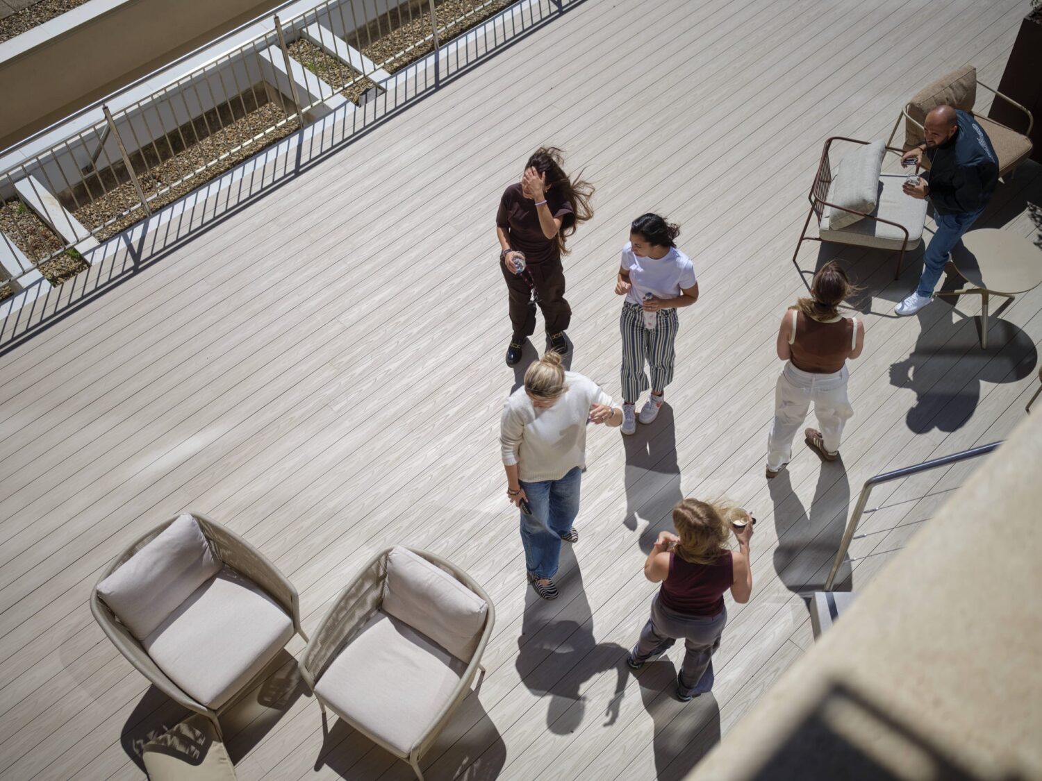 People stand and chat on a sunlit wooden terrace with white chairs and a railing in the background.