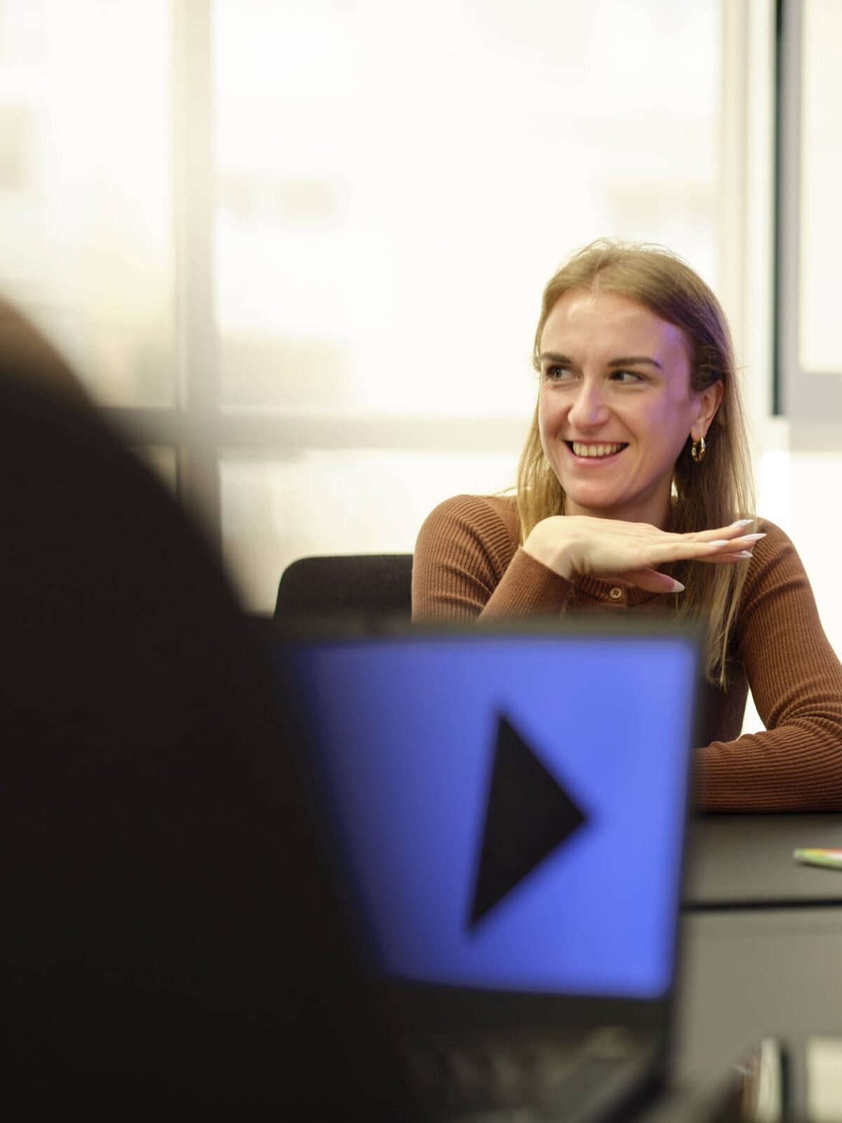 Smiling woman seated at a conference table with a laptop displaying a play button.
