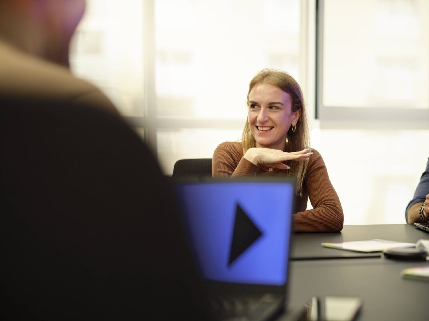 Smiling woman seated at a conference table with a laptop displaying a play button.