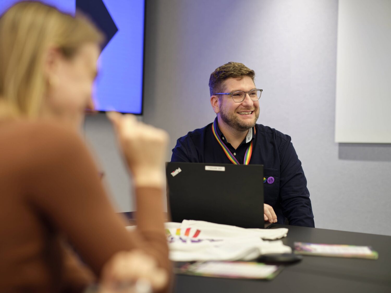 A man wearing glasses sits at a table with a laptop, smiling during a meeting.