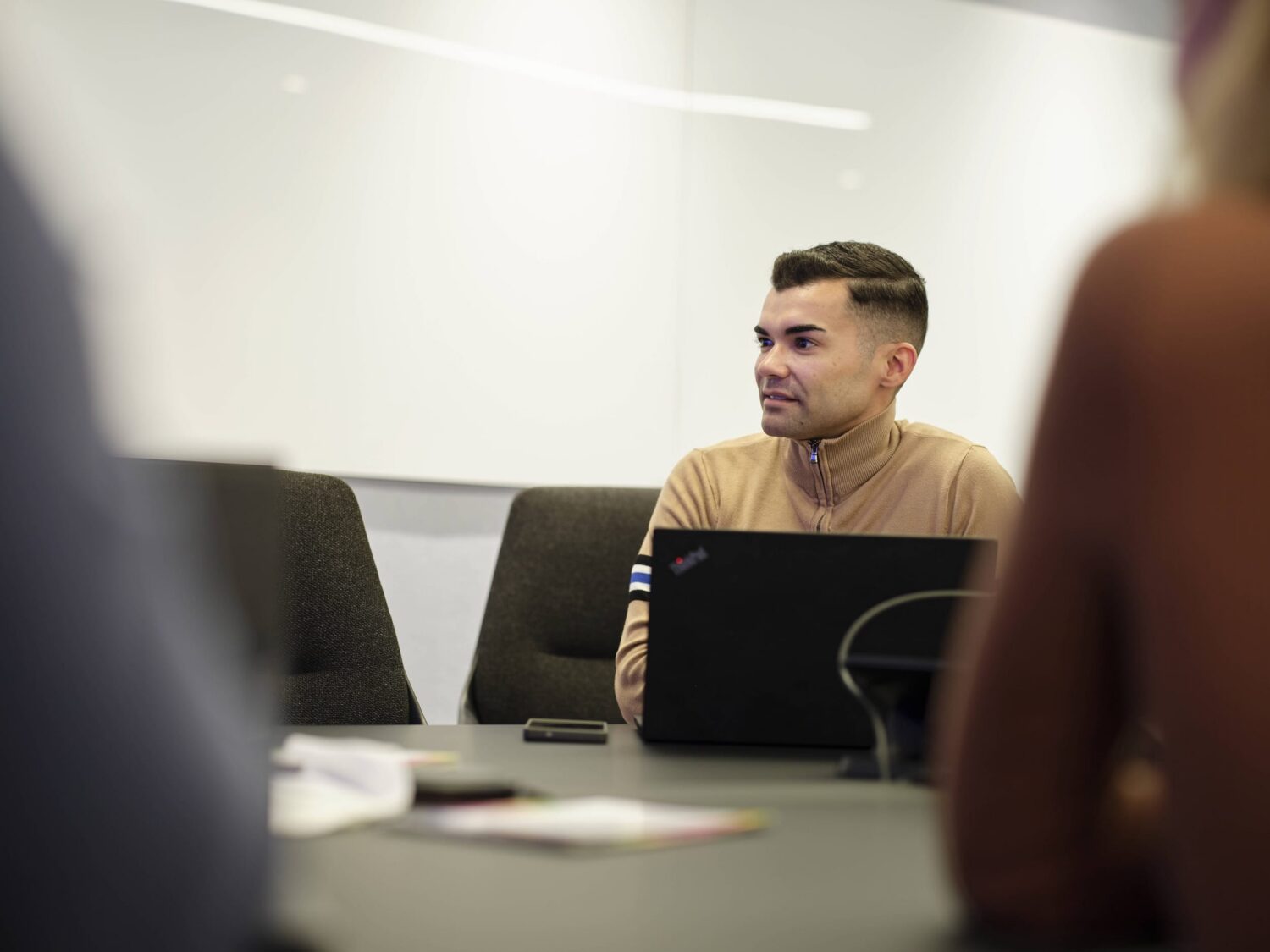 A man in a tan sweater sits at a conference table with a laptop in a meeting room.