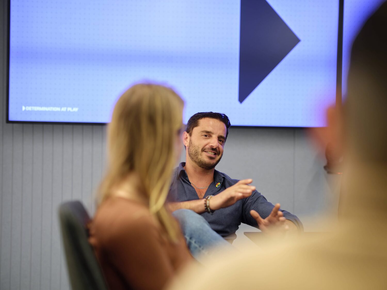 A man speaks during a panel discussion in a conference room, with blurred attendees in the foreground and a screen displaying a large play button in the background.
