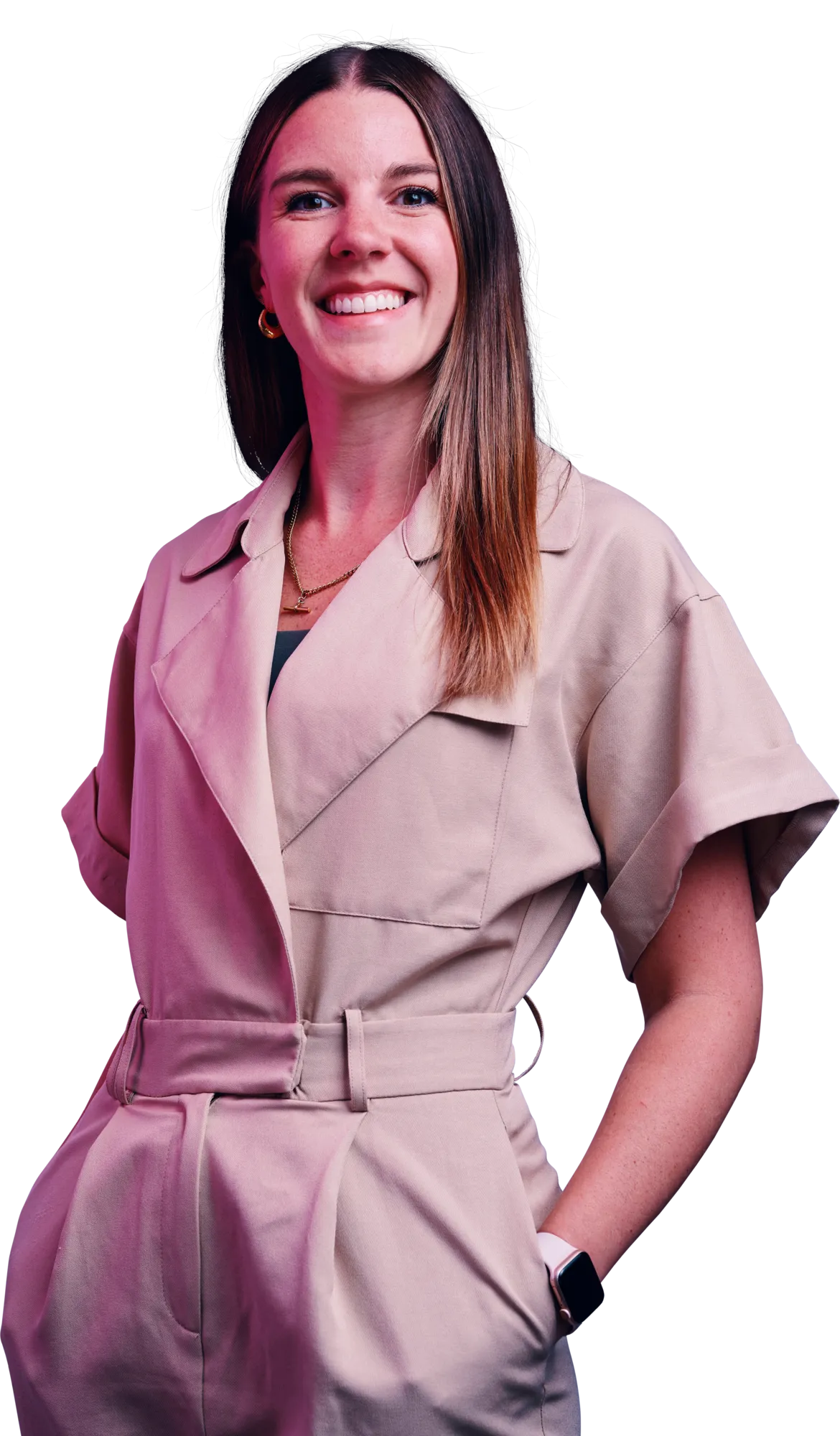 Smiling woman with long brown hair wearing a light beige belted jumpsuit, standing with one hand in her pocket.