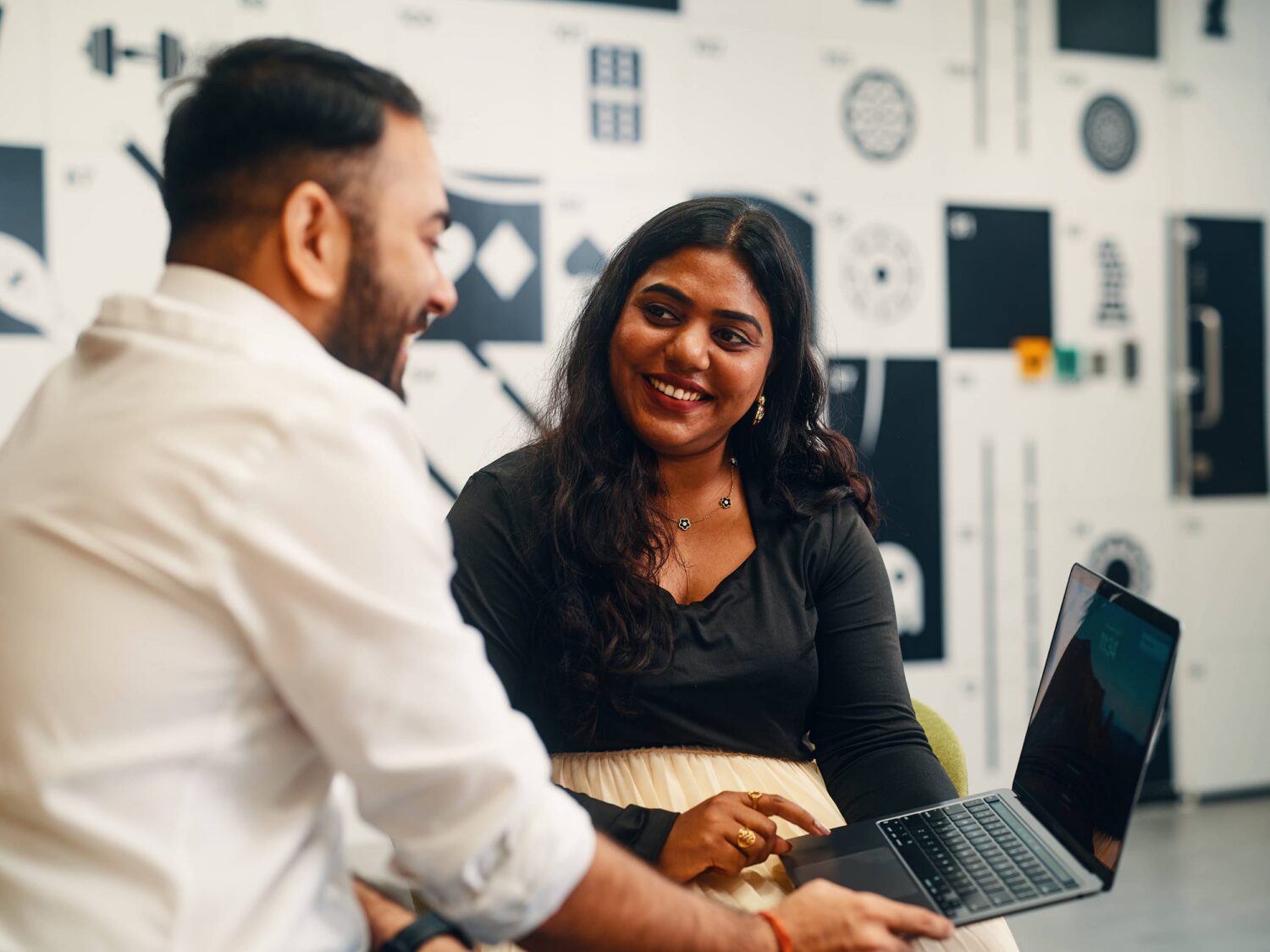 Two coworkers sit at a table in a bright modern office, smiling at an open laptop.