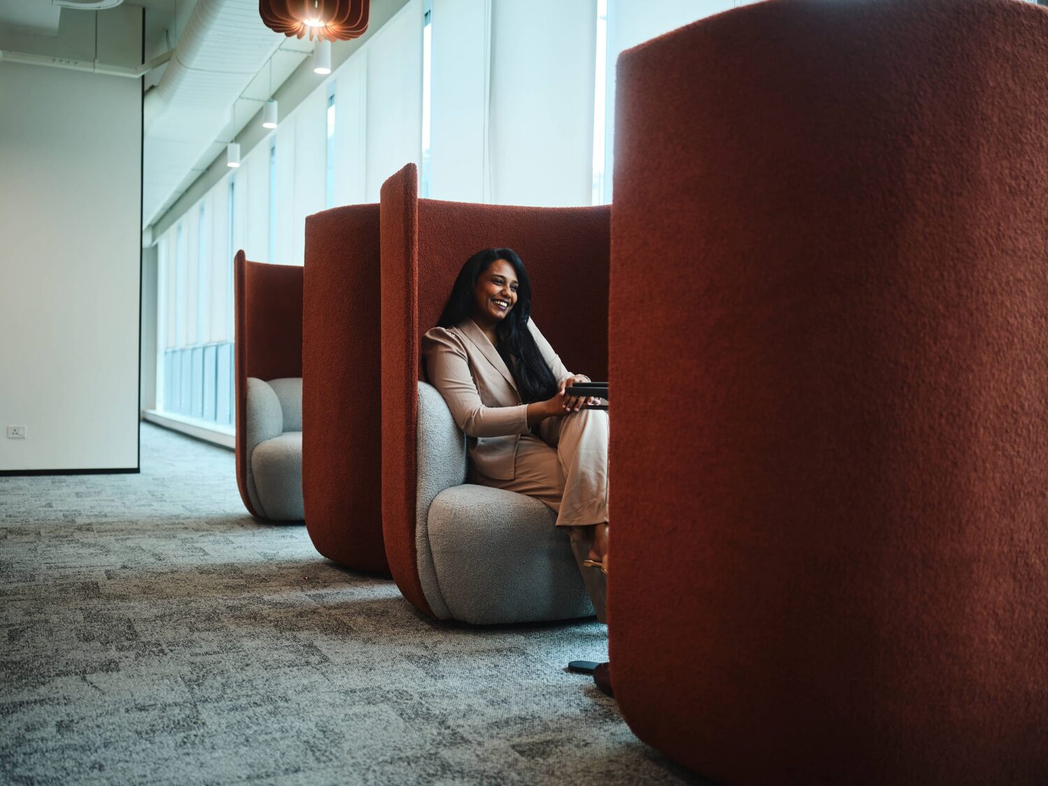 A woman sits inside a large curved red seating pod in a modern office lounge with large windows.