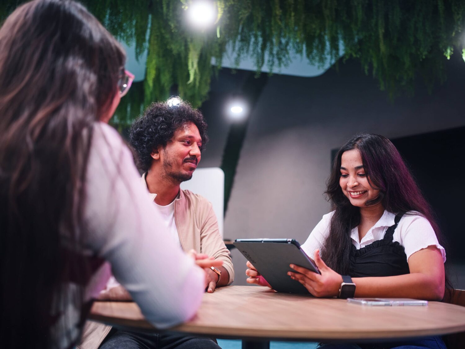 Three people sit around a wooden table indoors, smiling as one person holds a tablet.