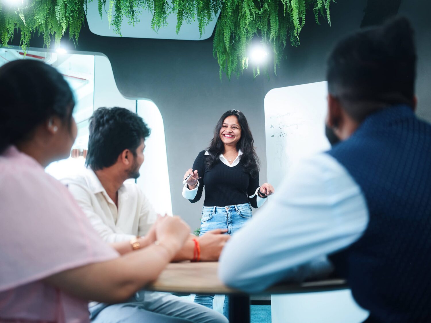 A woman presenter speaks to a small group seated around a table in a bright office with hanging greenery.