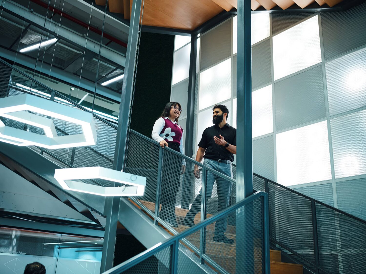 Two people stand on a glass-railed balcony in a modern building, talking near large windows and distinctive geometric light fixtures.