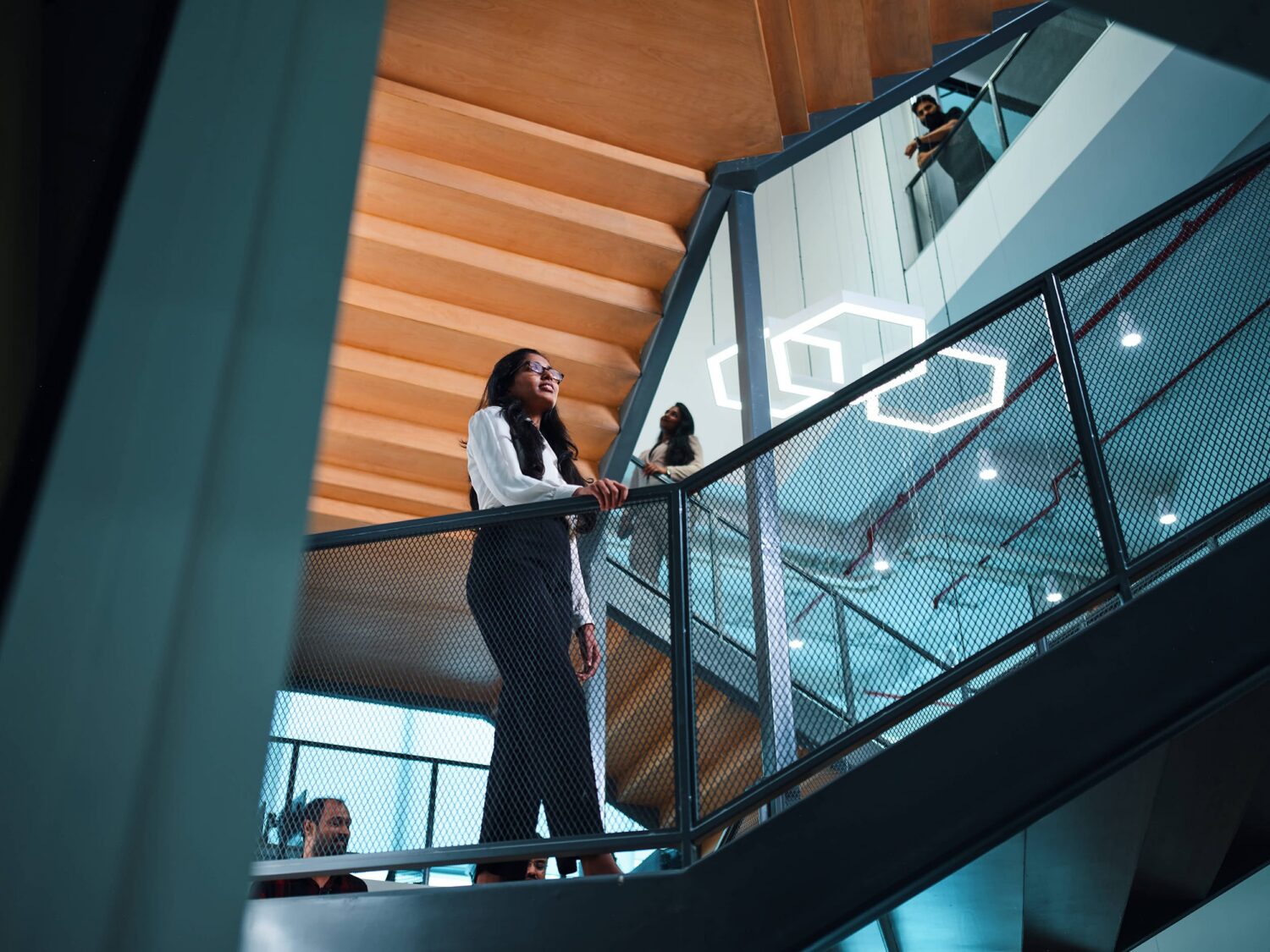 Two women stand on a glass-railed staircase inside a modern building with a hexagonal light fixture in the background.