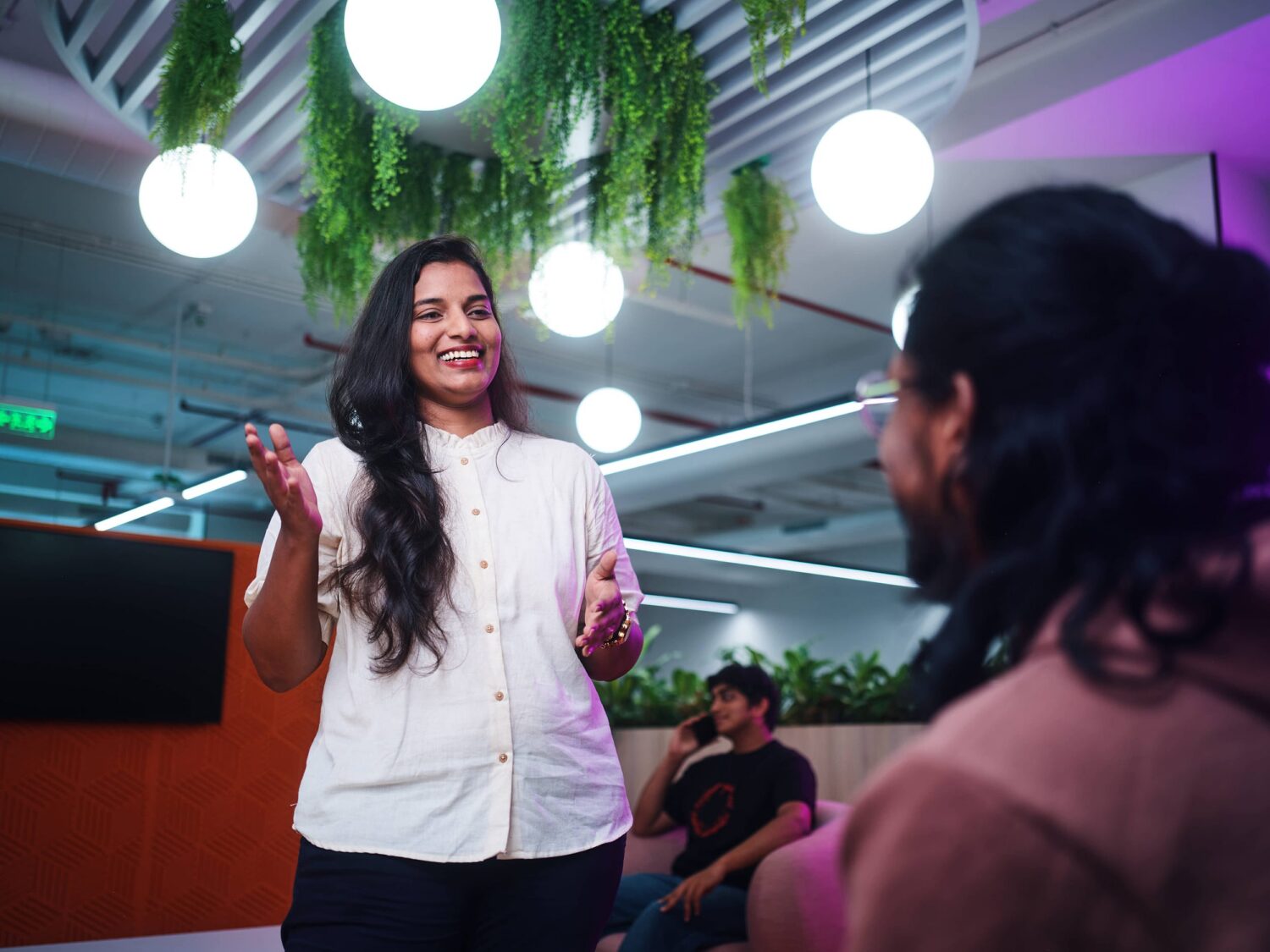 A woman with long dark hair wearing a white blouse stands and speaks to an audience in a modern room with hanging plants and round pendant lights.