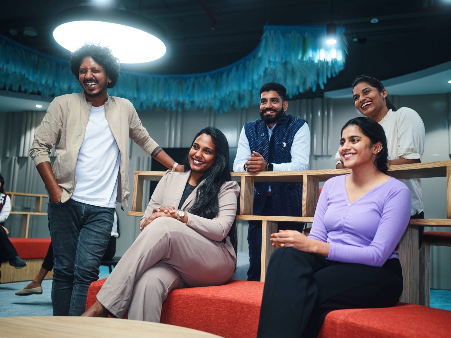 Six casually dressed people smiling and chatting in a modern office lounge with red seating.