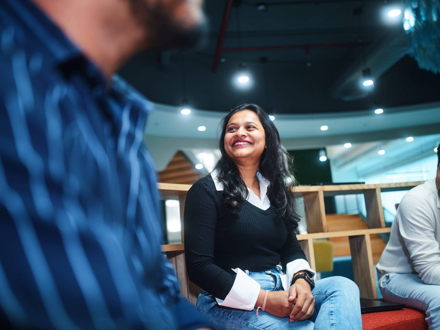 A smiling woman with long dark hair wearing a black top and jeans sits in a modern indoor space with others nearby.