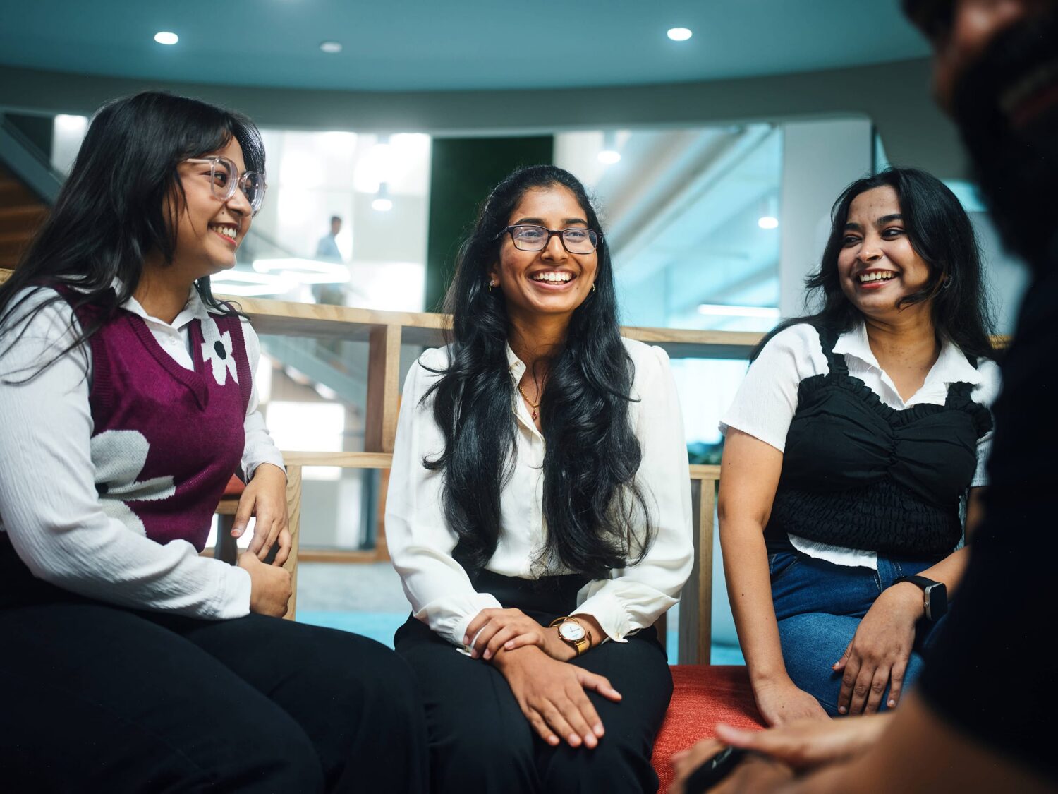 Three smiling women sit together in a modern, well-lit office or classroom setting.