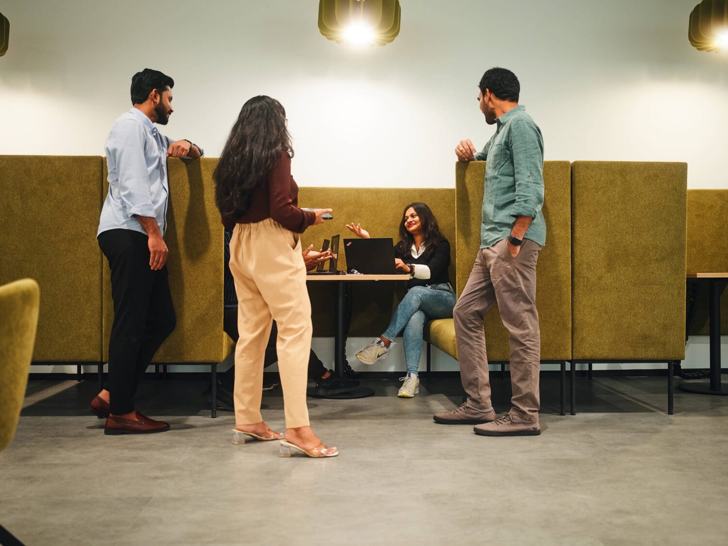 Six people in a casual cafe, with four standing around a table while two sit and work on a laptop.