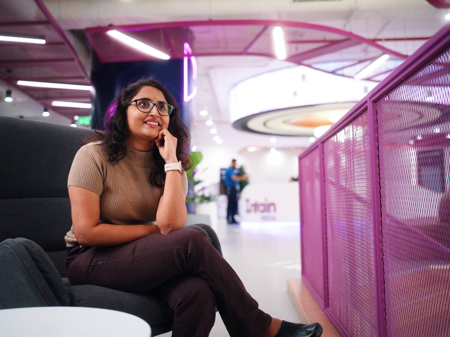 A smiling woman wearing glasses and a brown top sits on a white chair in a bright modern coworking space with purple railings.