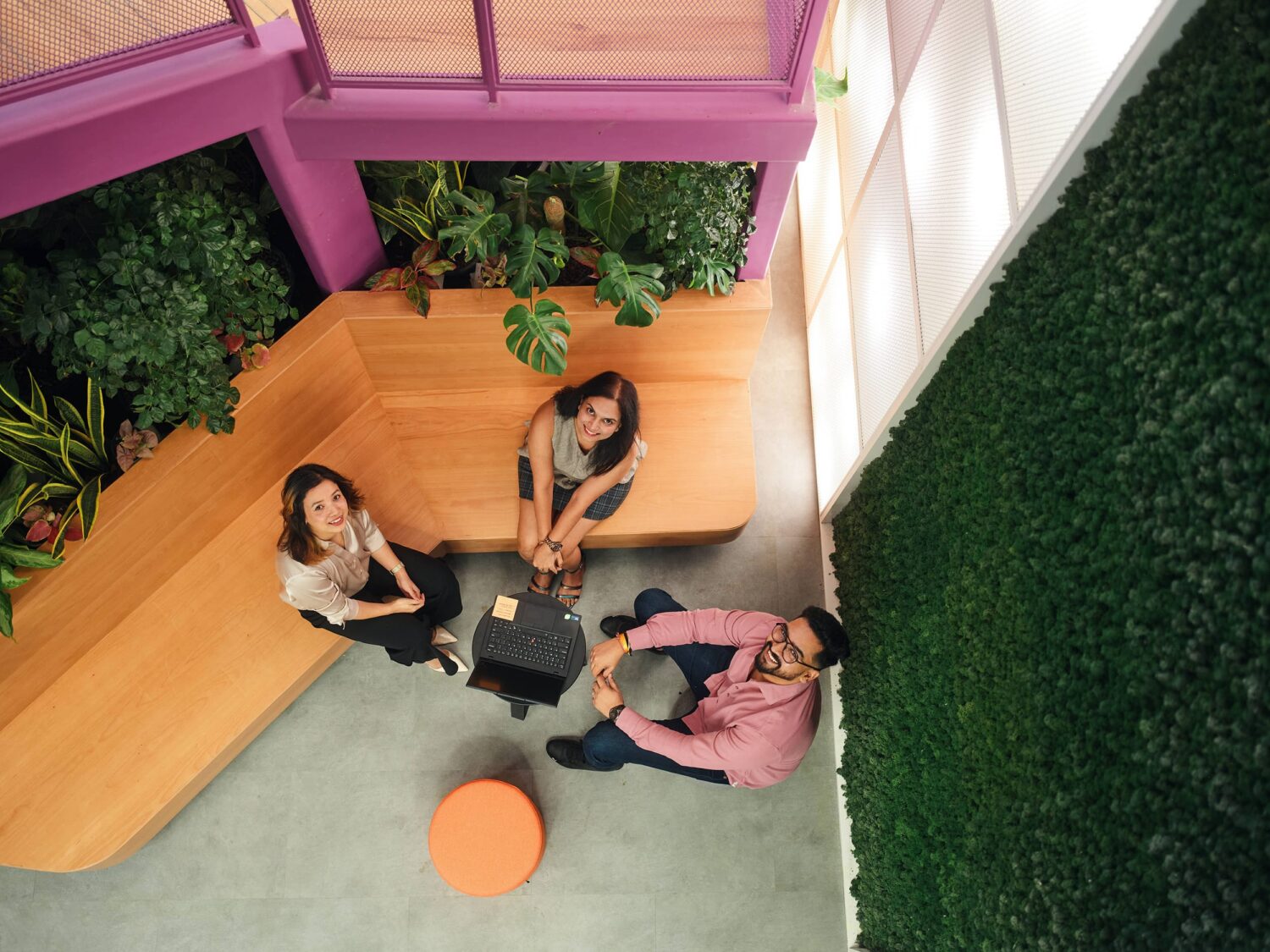 People sit around a small square table in a bright indoor space with purple beams, wooden seating, and a green plant wall, viewed from above.