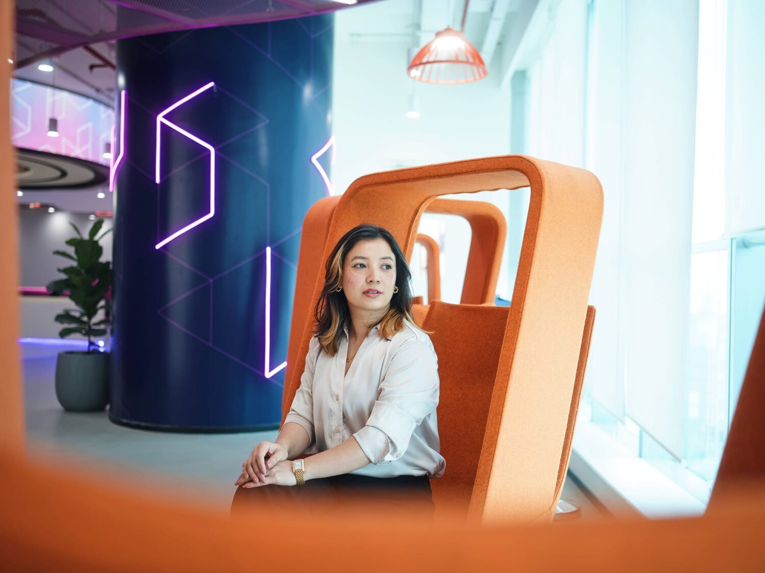 A woman sits in an orange curved chair in a bright, futuristic interior with purple neon wall panels.