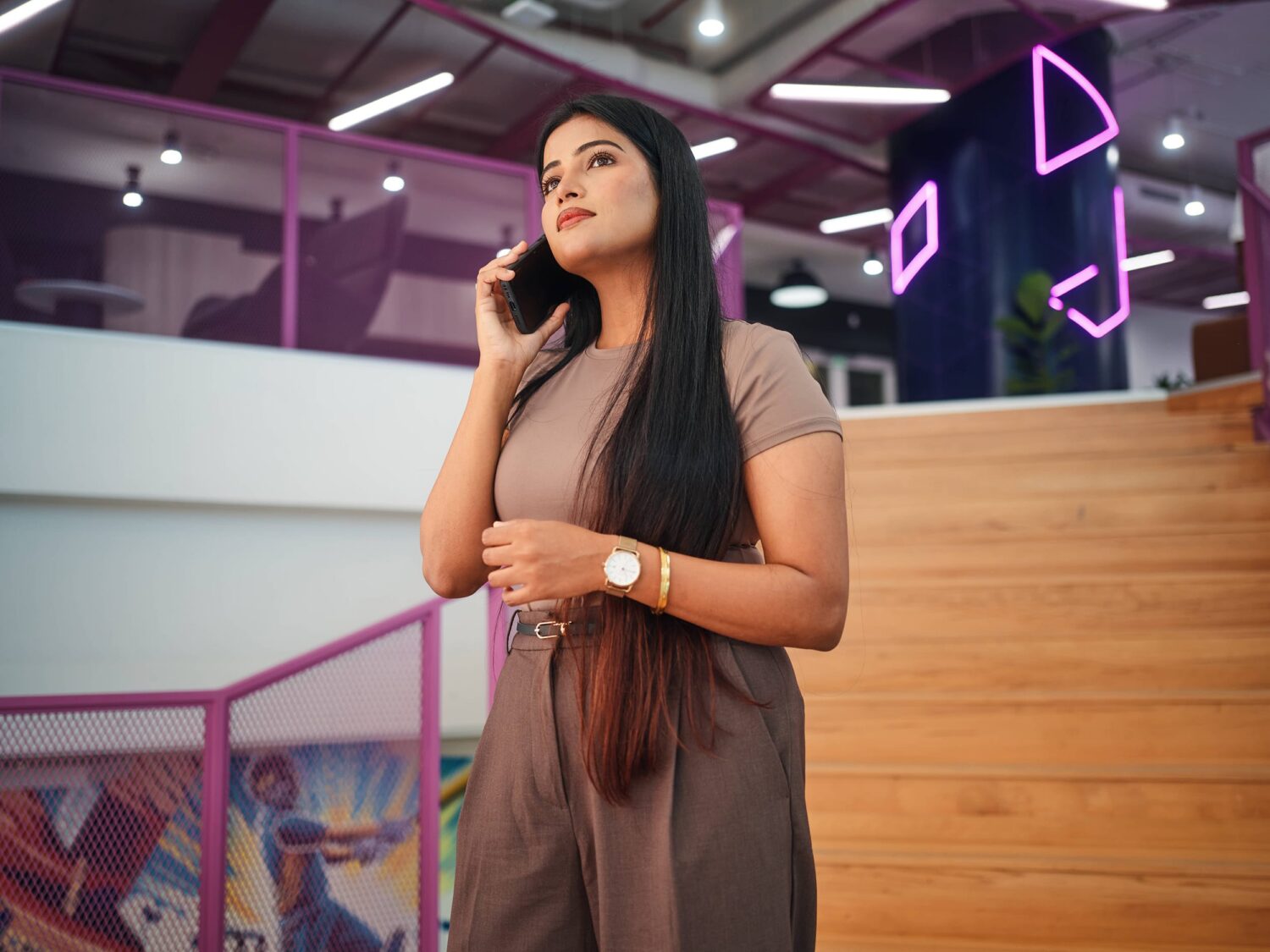 A woman with long dark hair wearing a taupe outfit talks on her cell phone inside a neon-lit space with a wooden staircase.