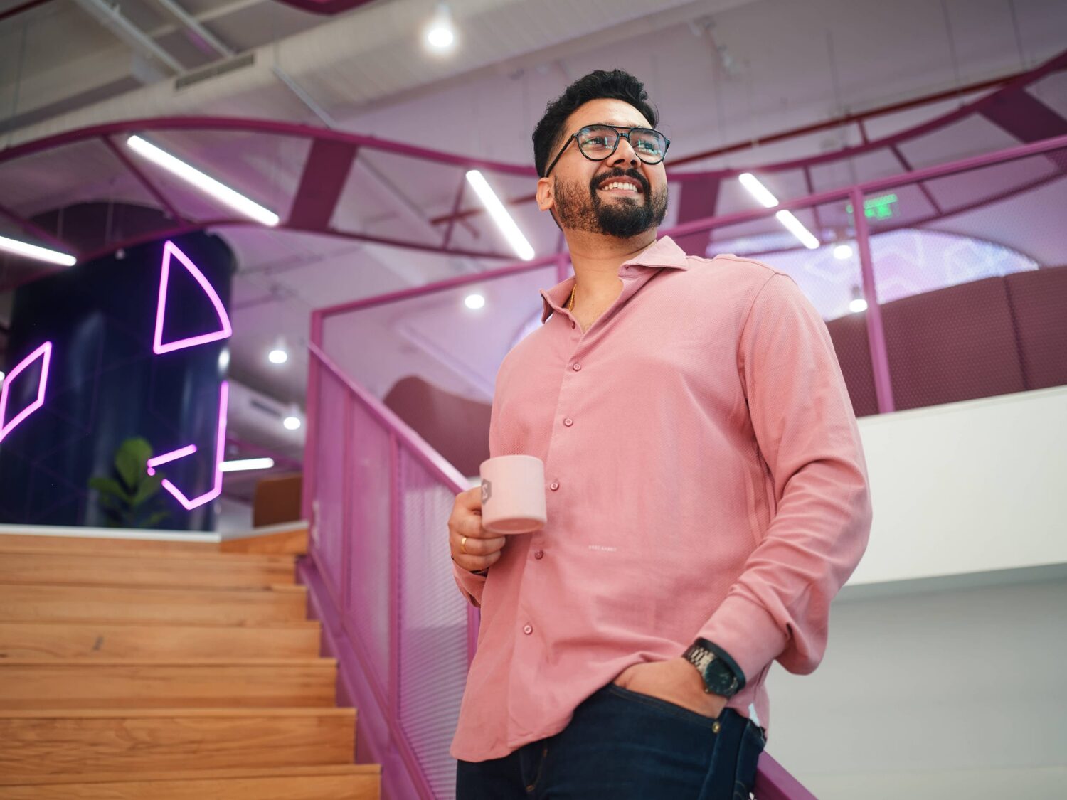 A man in a pink shirt and glasses holds a cup while standing in a colorful, modern interior with purple railings and neon lights.