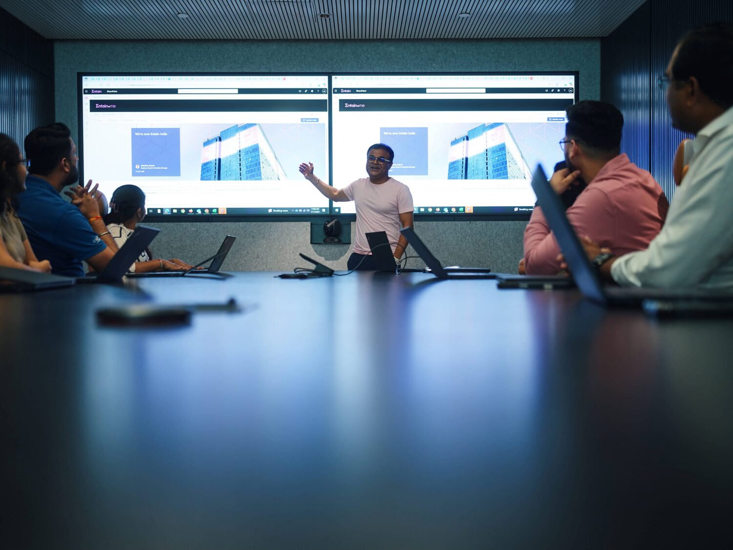 A presenter leads a meeting in a conference room as attendees with laptops watch two large screens displaying slides and charts.