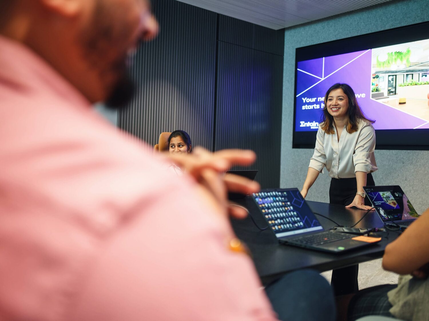 A presenter stands at the front of a meeting room with a laptop on the table and a large screen displaying slides behind her.