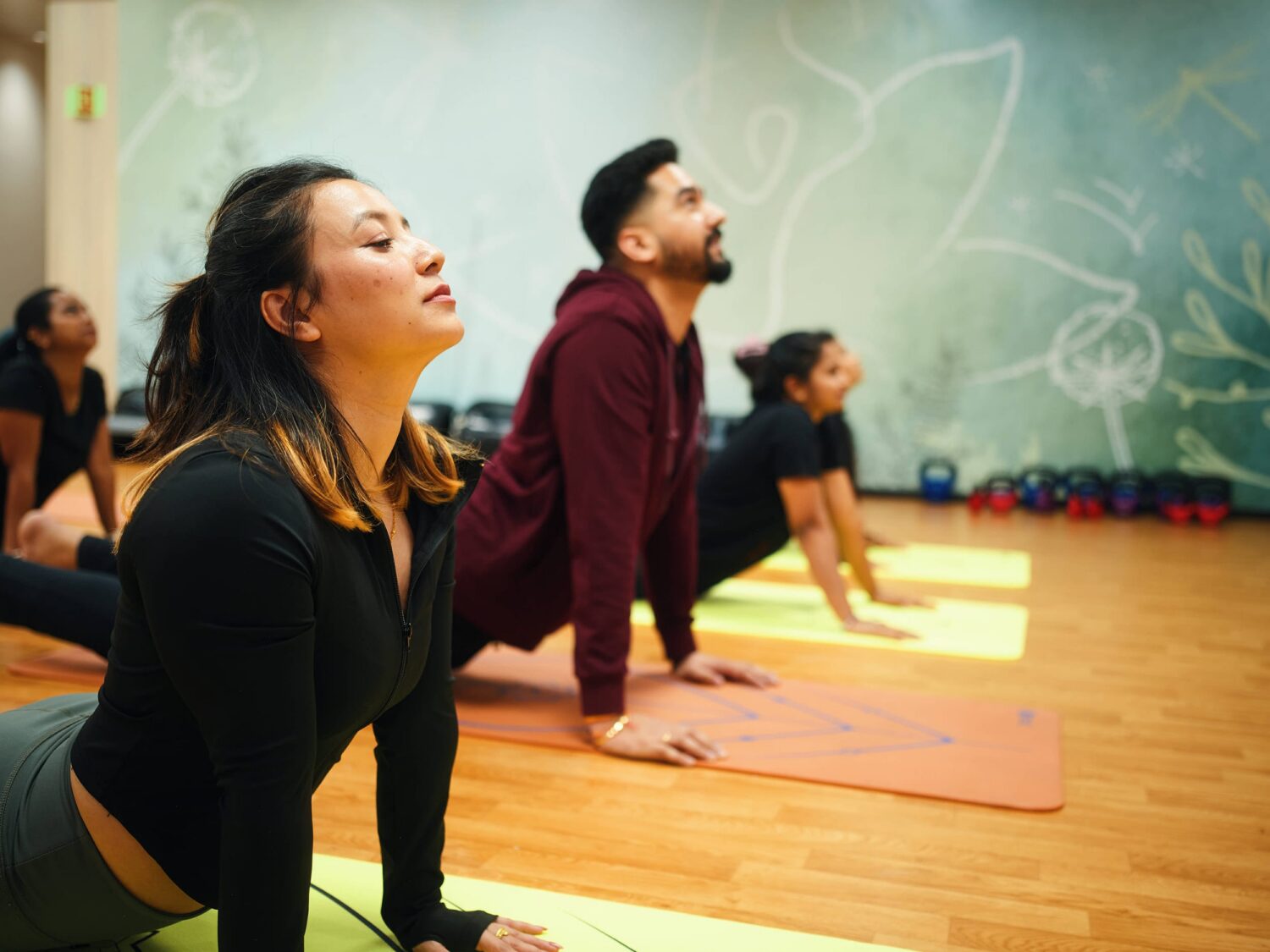 A group of people practicing yoga on colorful mats in a studio with a chalk-drawn leafy mural.