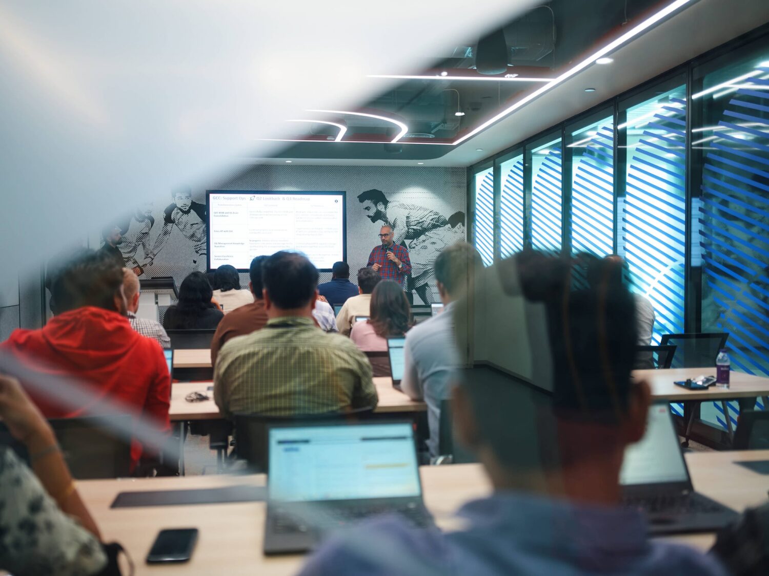 A group of people seated with laptops in a conference room watch a presenter at the front near a projector screen.