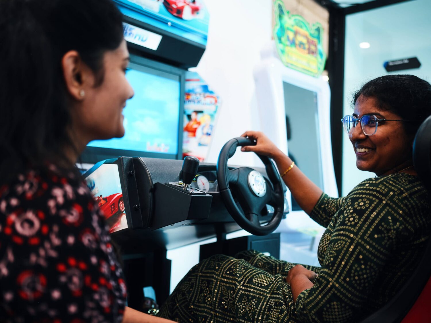 Two women sit in a driving simulator at an arcade, one driving with a steering wheel and smiling.