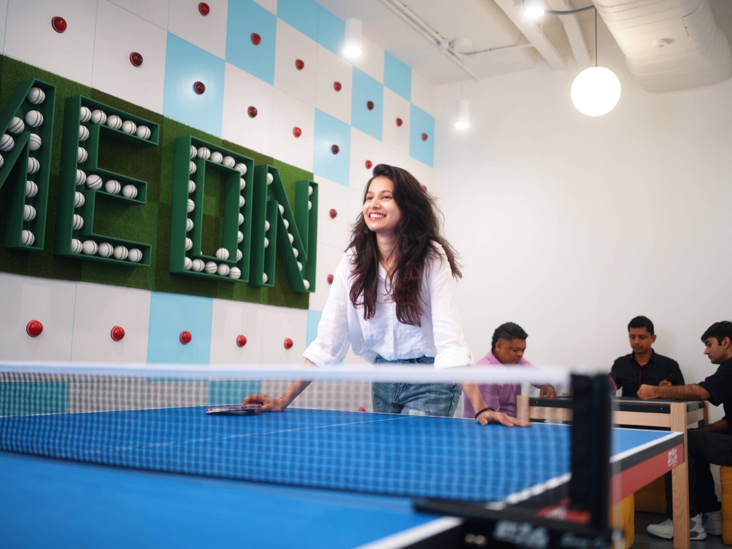 A smiling young woman plays table tennis indoors at a blue table, with others seated in the background.