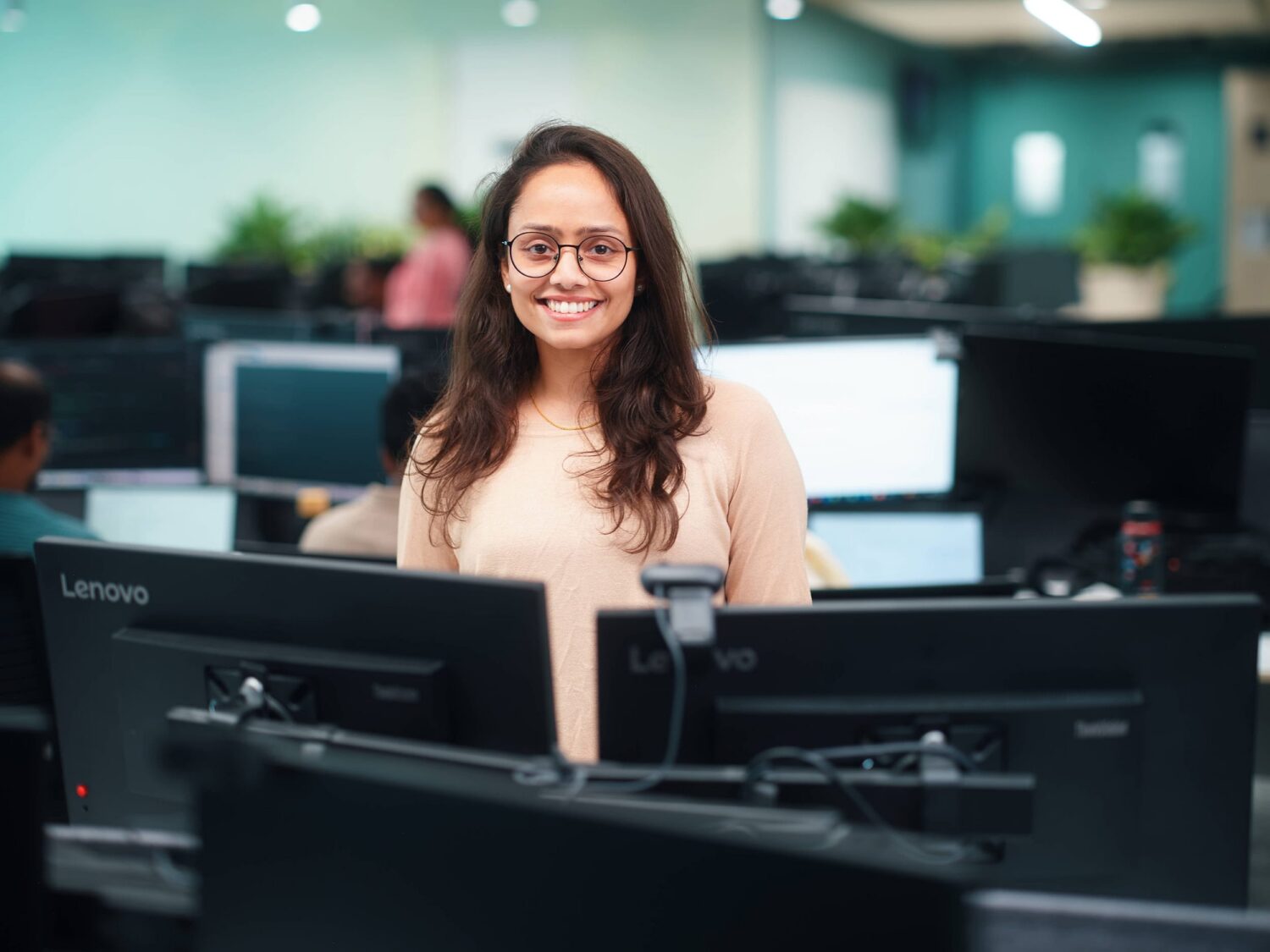 Smiling woman with glasses sits at a desk in a busy office surrounded by multiple computer monitors.