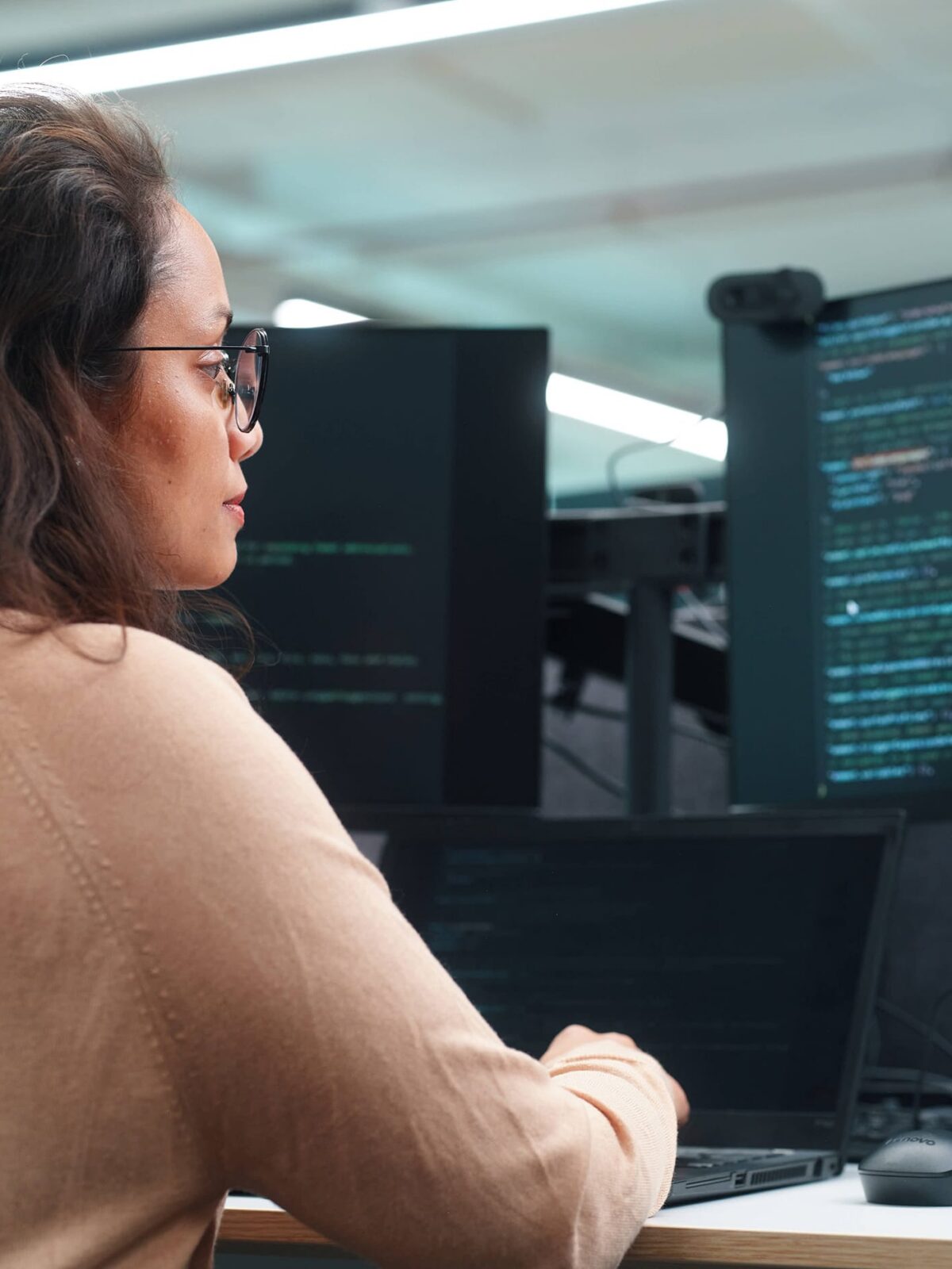 A woman with glasses sits at a desk in front of multiple monitors displaying code.