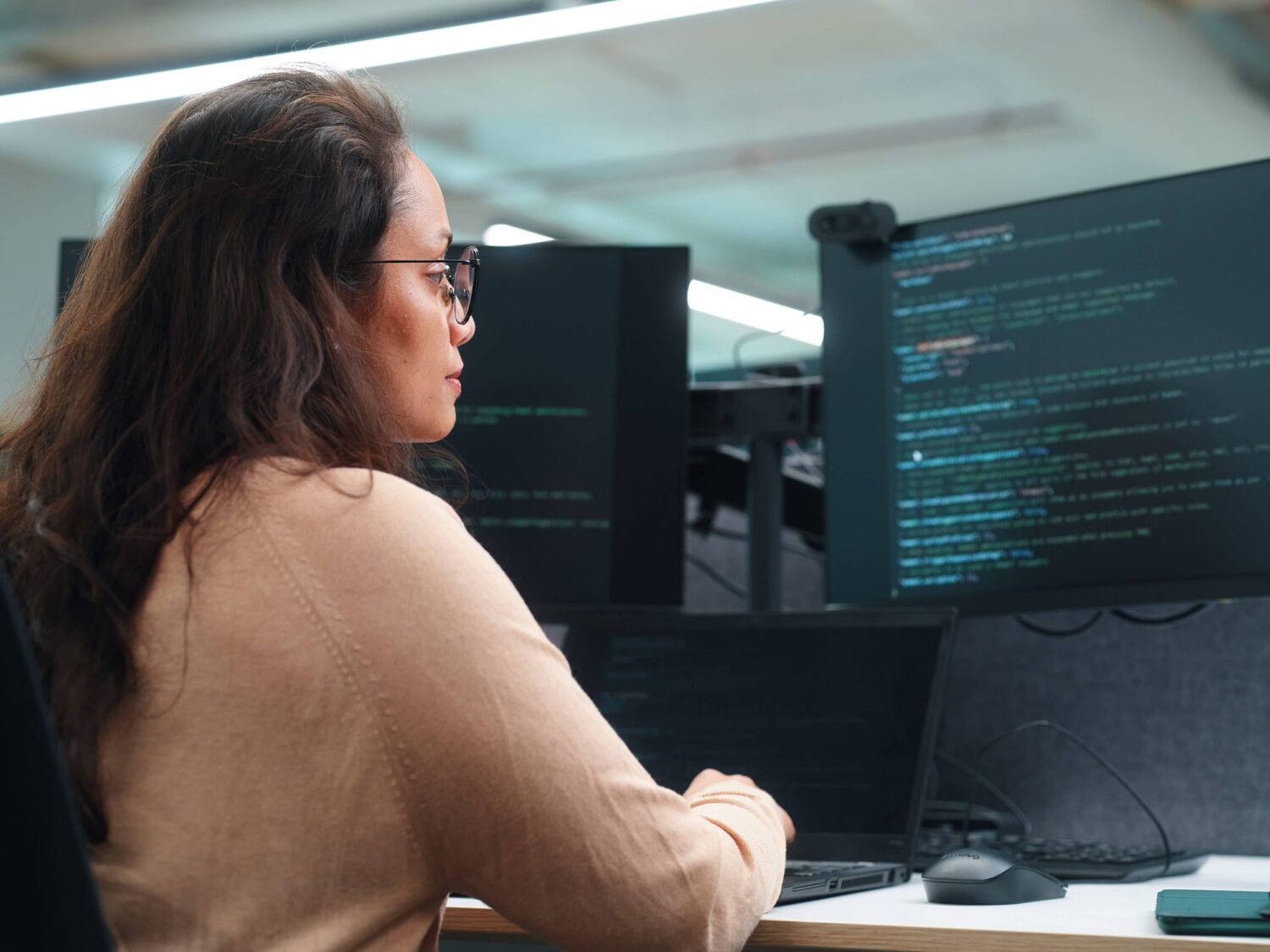 A woman with glasses sits at a desk in front of multiple monitors displaying code.