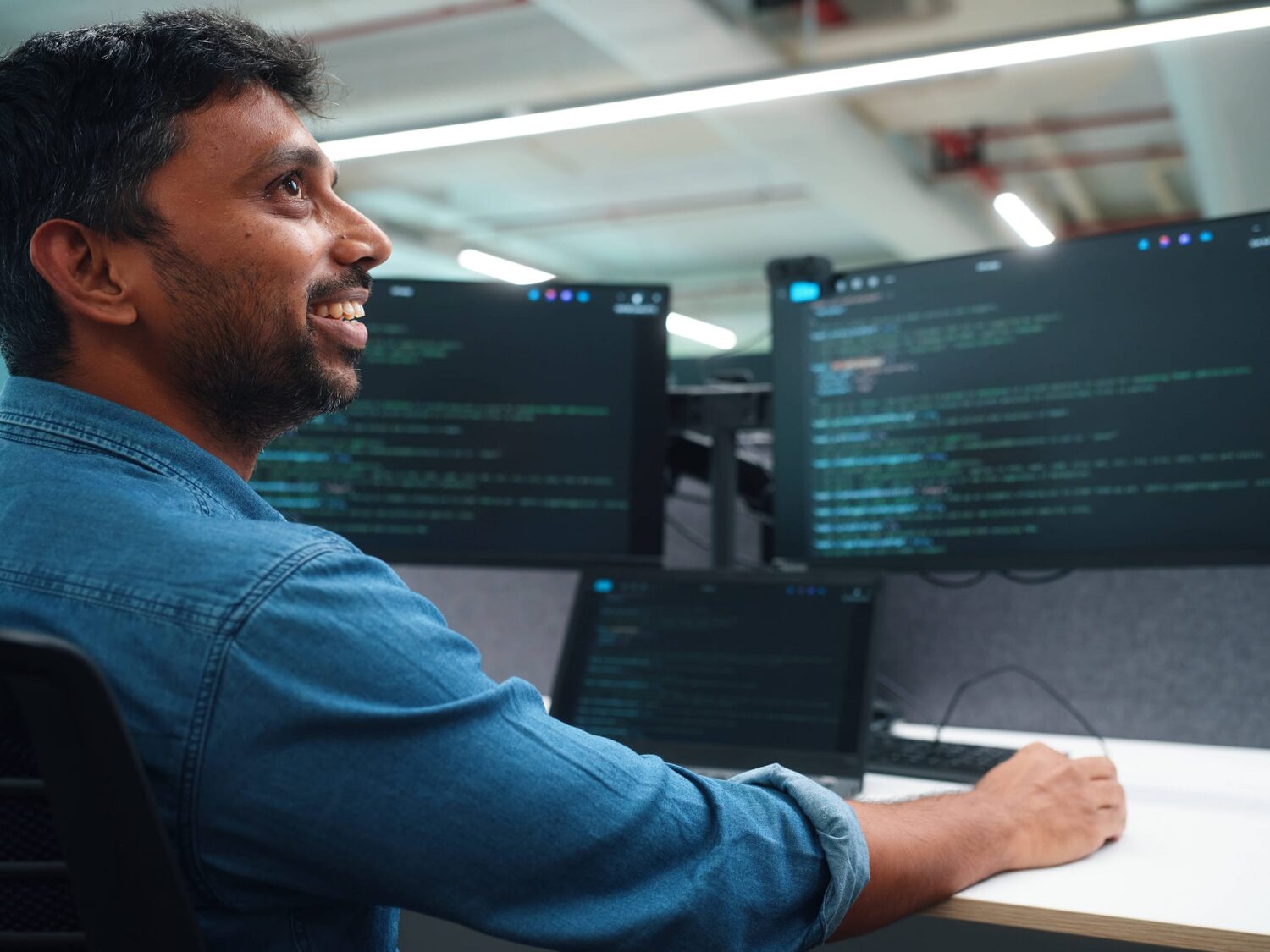 A man in a blue shirt sits at a desk coding on multiple monitors, smiling.