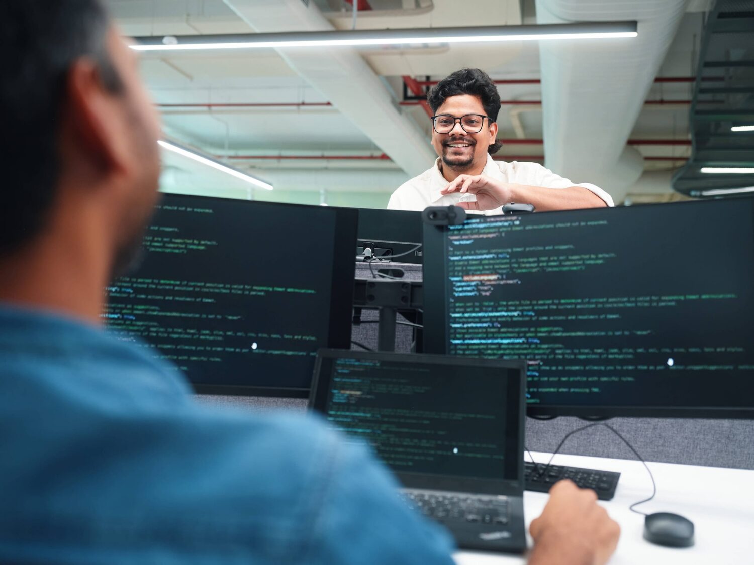 Two software developers collaborate in an office, with multiple monitors displaying code.